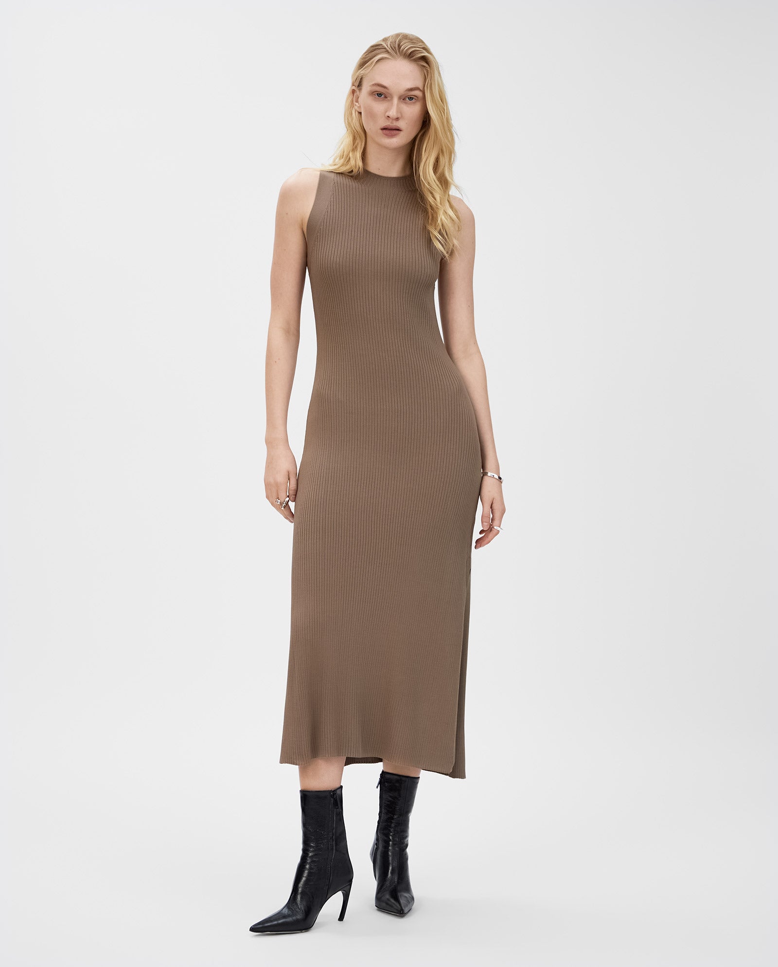 A woman wears the IVY OAK KLAUDIA Dress, a sleeveless, fitted taupe rib-knit midi dress, styled with black heeled boots against a white background.