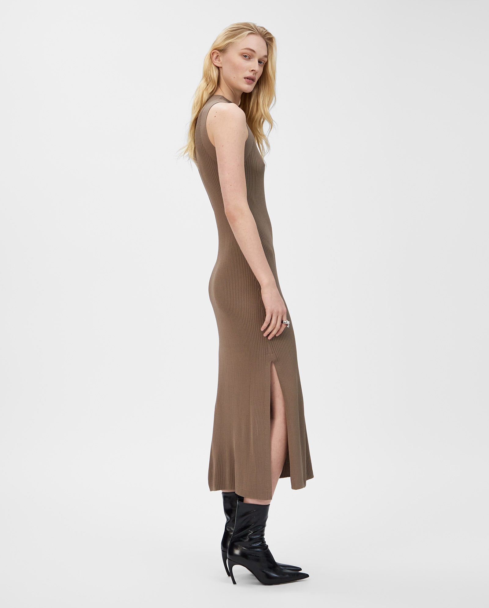 A woman stands sideways wearing the IVY OAK KLAUDIA sleeveless brown ribbed knit dress with a side slit, paired with black boots.