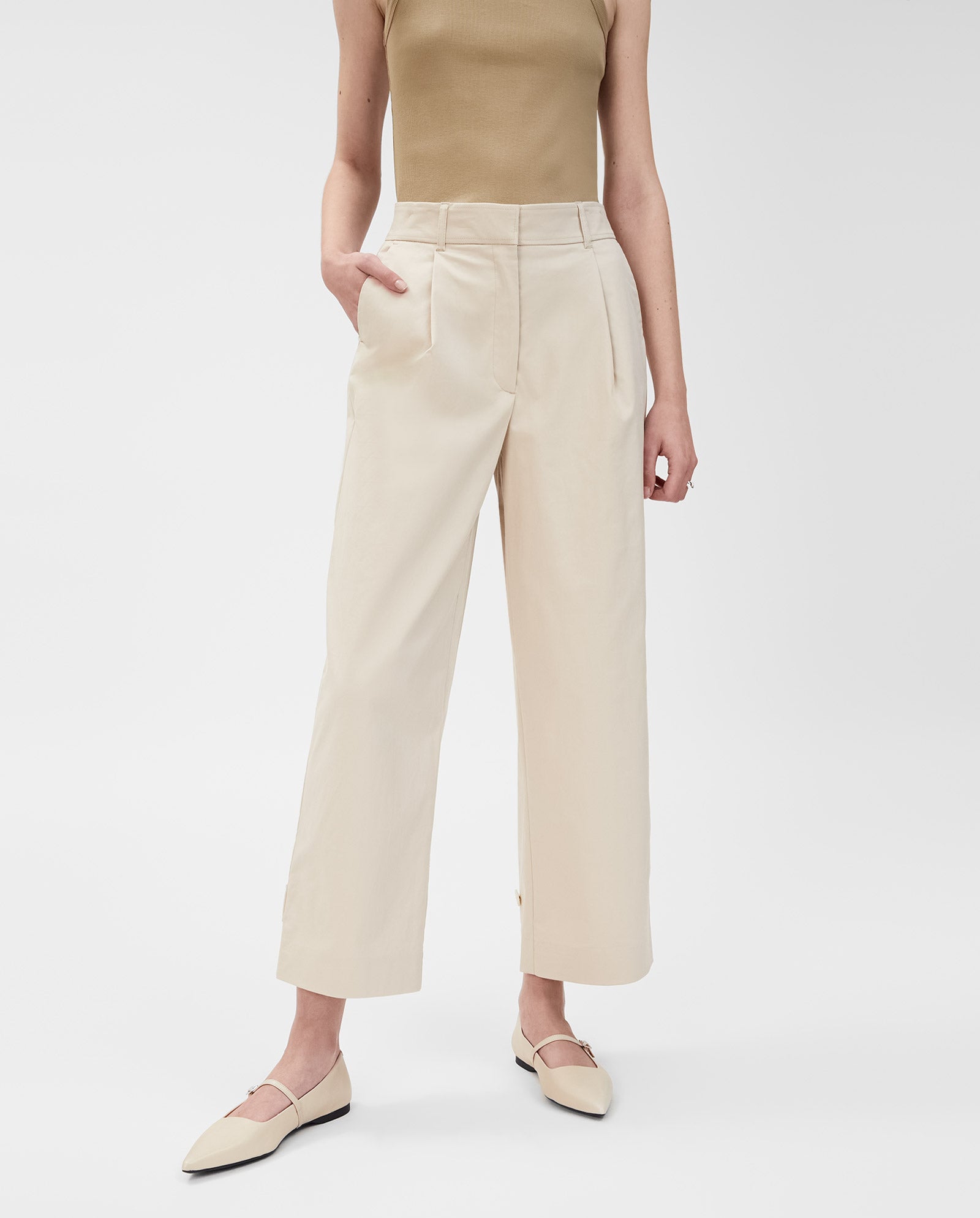 A woman stands against a white background wearing IVY OAKs PERSEPHONE Trousers—high-waisted beige wide-leg pants—paired with a tan sleeveless top and cream flats.