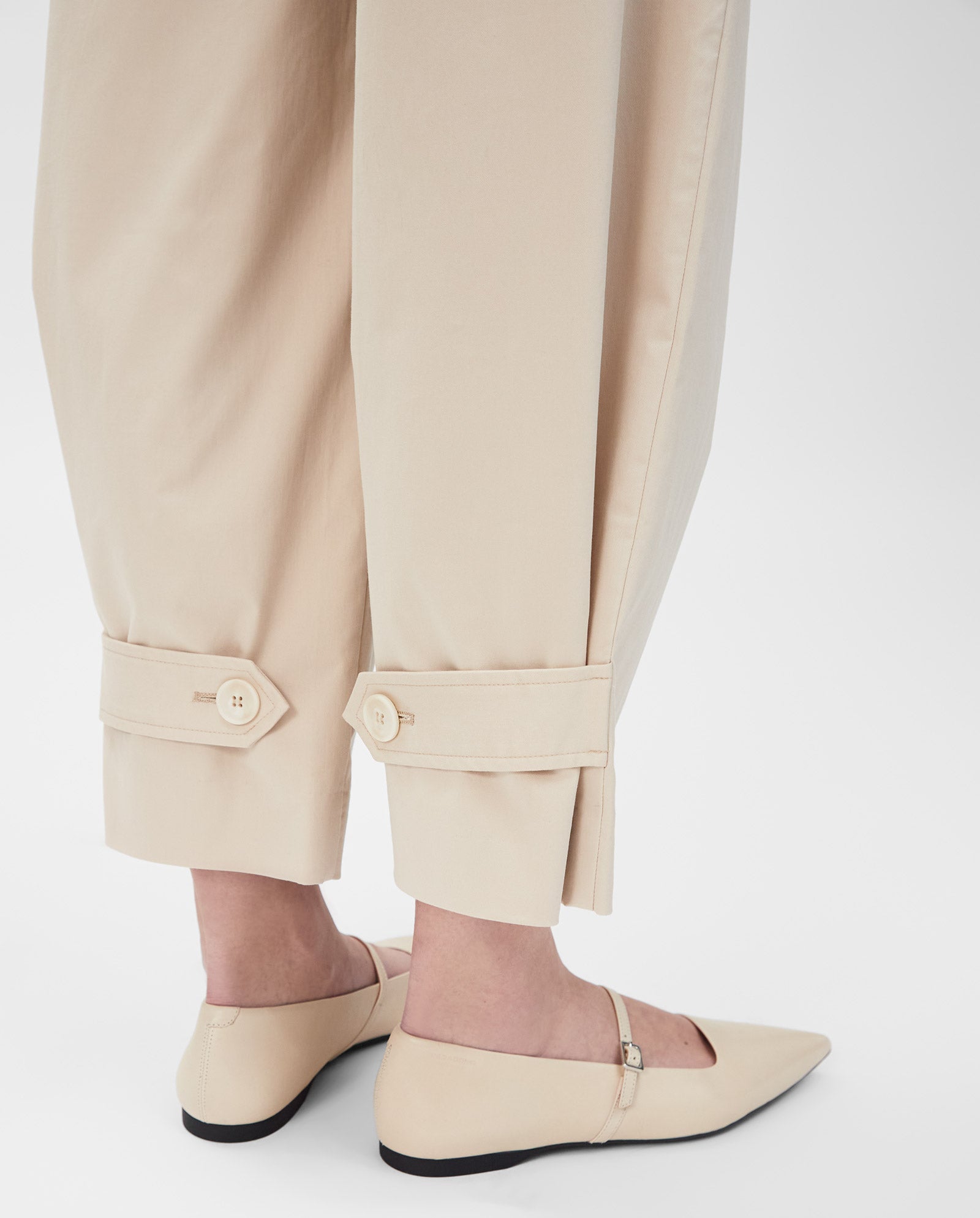 Close-up of IVY OAKs PERSEPHONE Trousers—beige, high-waisted, cuffed with button details—styled with beige pointed-toe flats for a sleek, tapered look.