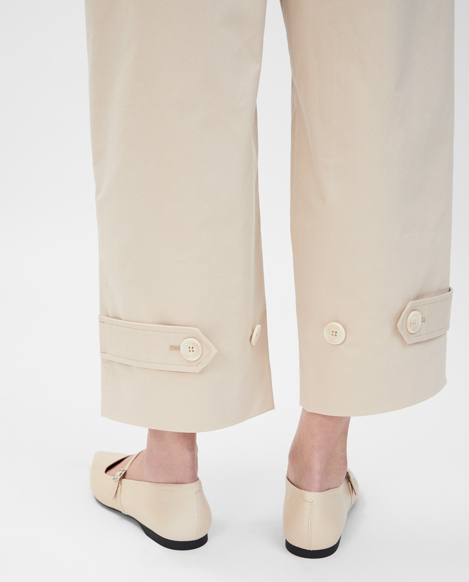 Model wearing IVY OAK PERSEPHONE high-waisted beige wide-leg trousers with buttoned cuffs and beige flats, shown from the calves down.