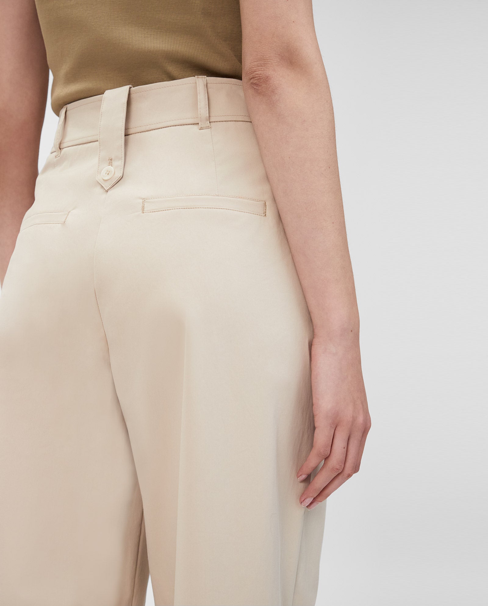 Model wearing IVY OAK’s PERSEPHONE Trousers in beige, styled high-waisted with a brown top. Seen from the back with one arm relaxed, the chic tapered silhouette is highlighted.