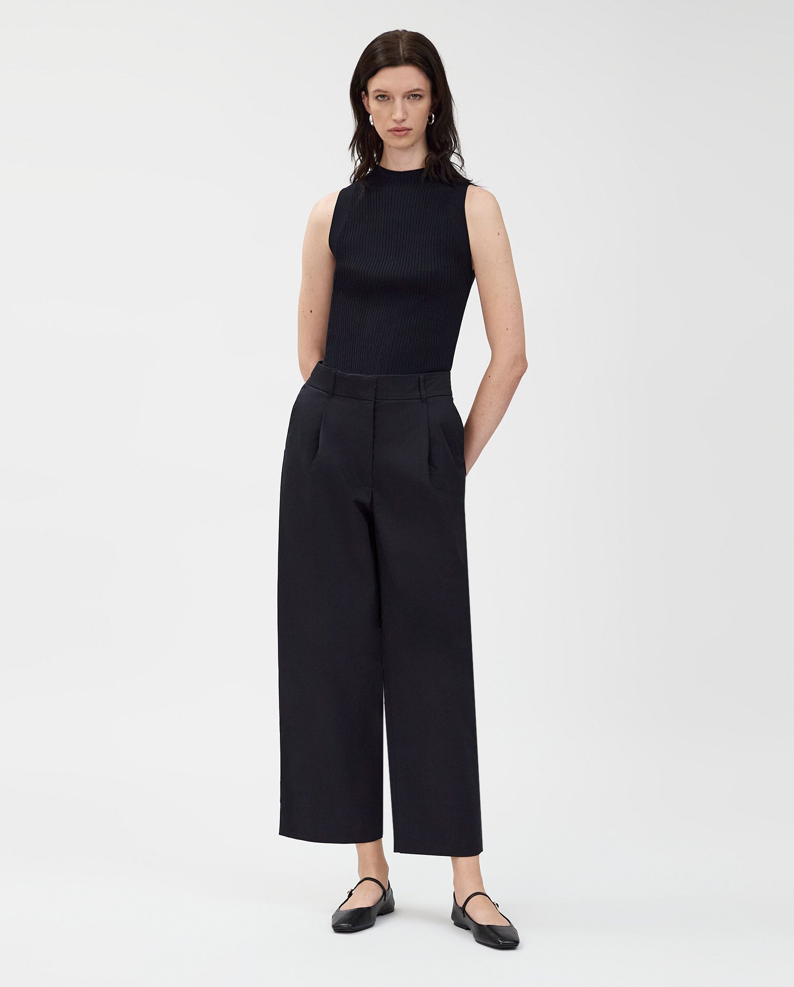 A woman stands against a white background wearing IVY OAKs PERSEPHONE Trousers, featuring a high waist and adjustable hem, paired with a sleeveless black top and black flats.