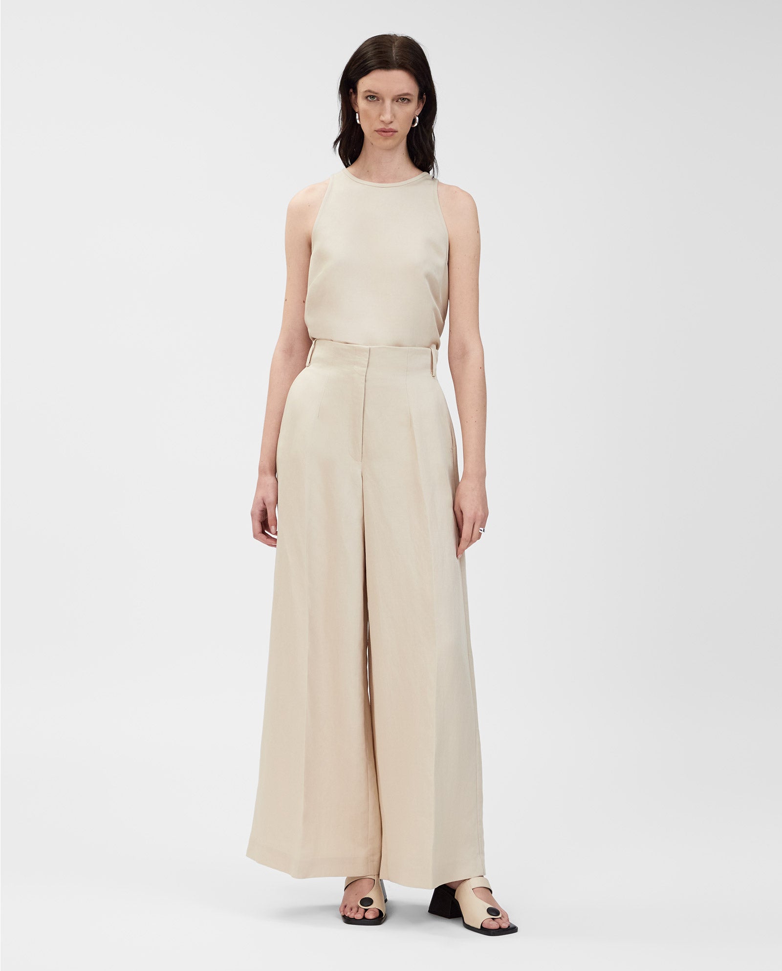 A woman models IVY OAK’s PIERA Trousers, featuring a wide-leg silhouette in beige, paired with a matching sleeveless top and set against a plain white background.