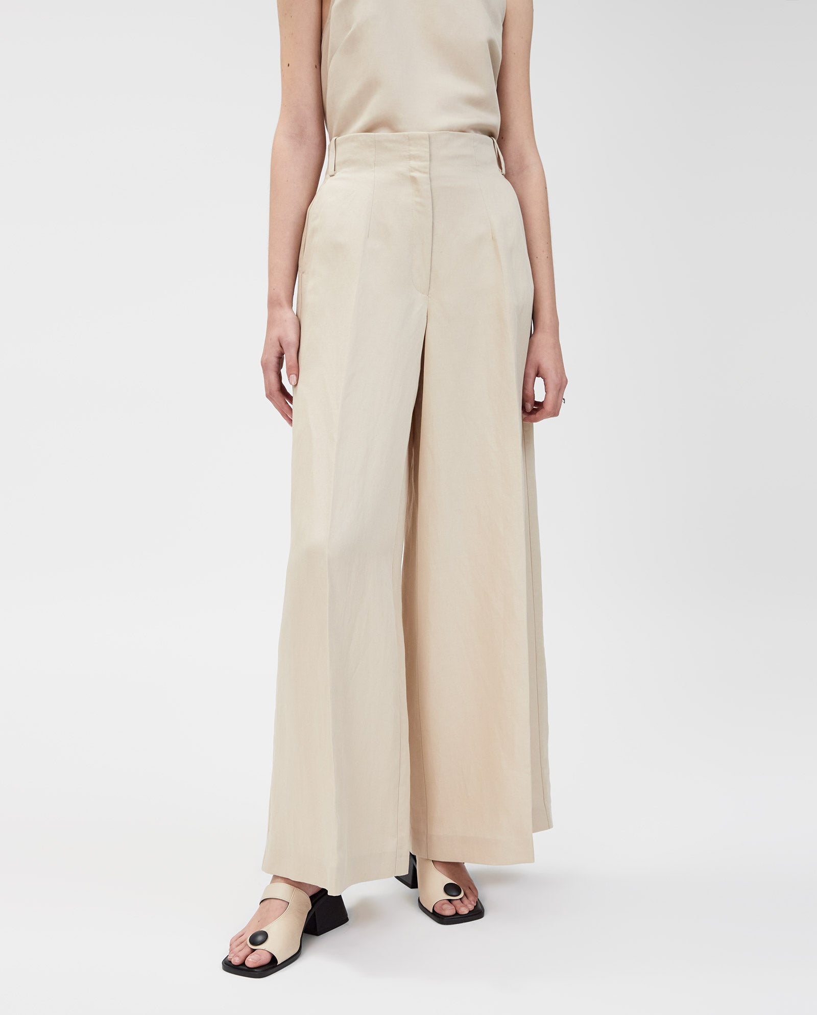 Model wears IVY OAK’s PIERA Trousers in beige with a high waist and wide-leg cut, paired with a sleeveless beige top and open-toe heeled sandals, set against a white background.