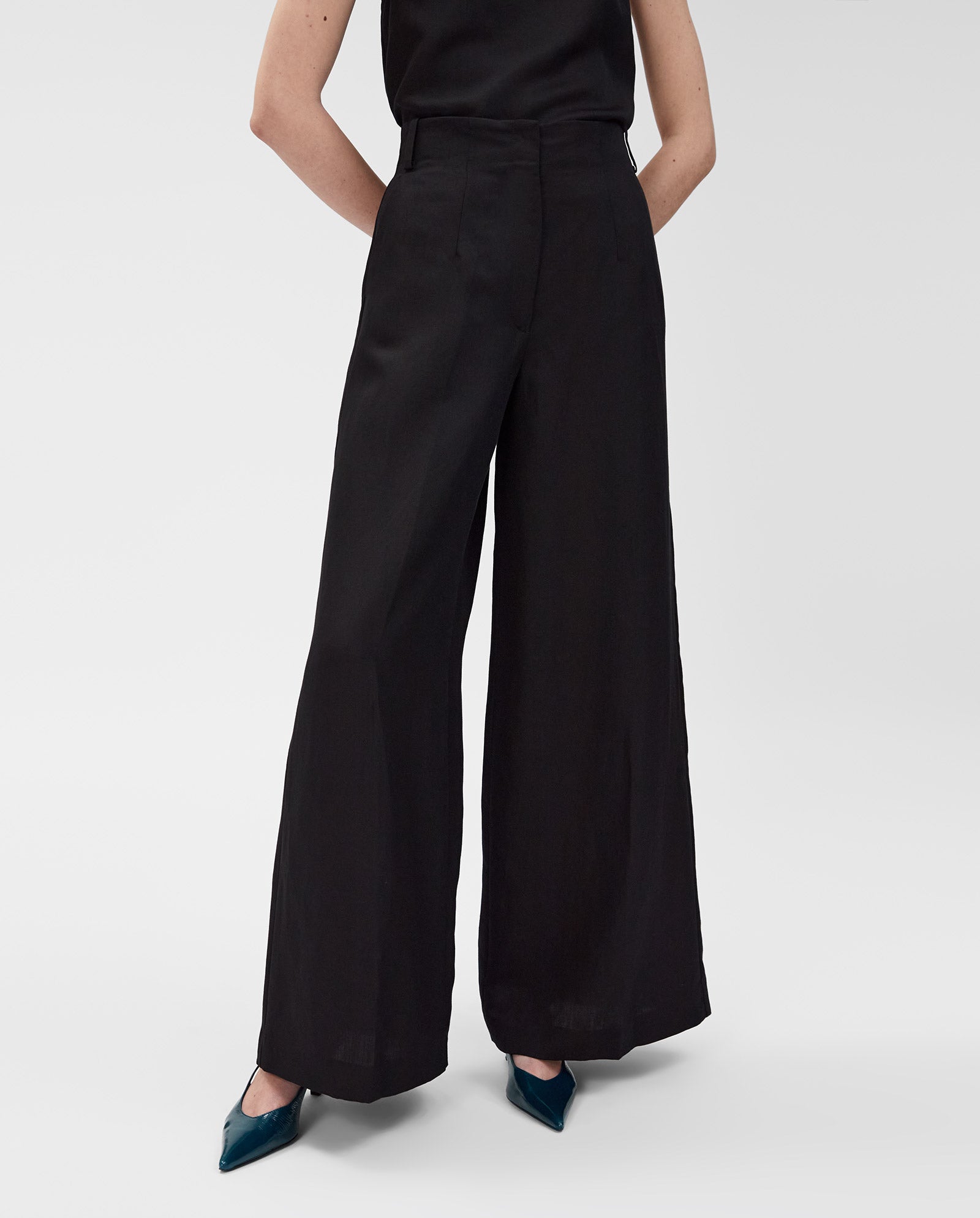 A person wearing IVY OAKs PIERA Trousers, featuring a high waist and wide legs in black linen blend, paired with teal pointed shoes, stands with hands behind their back.