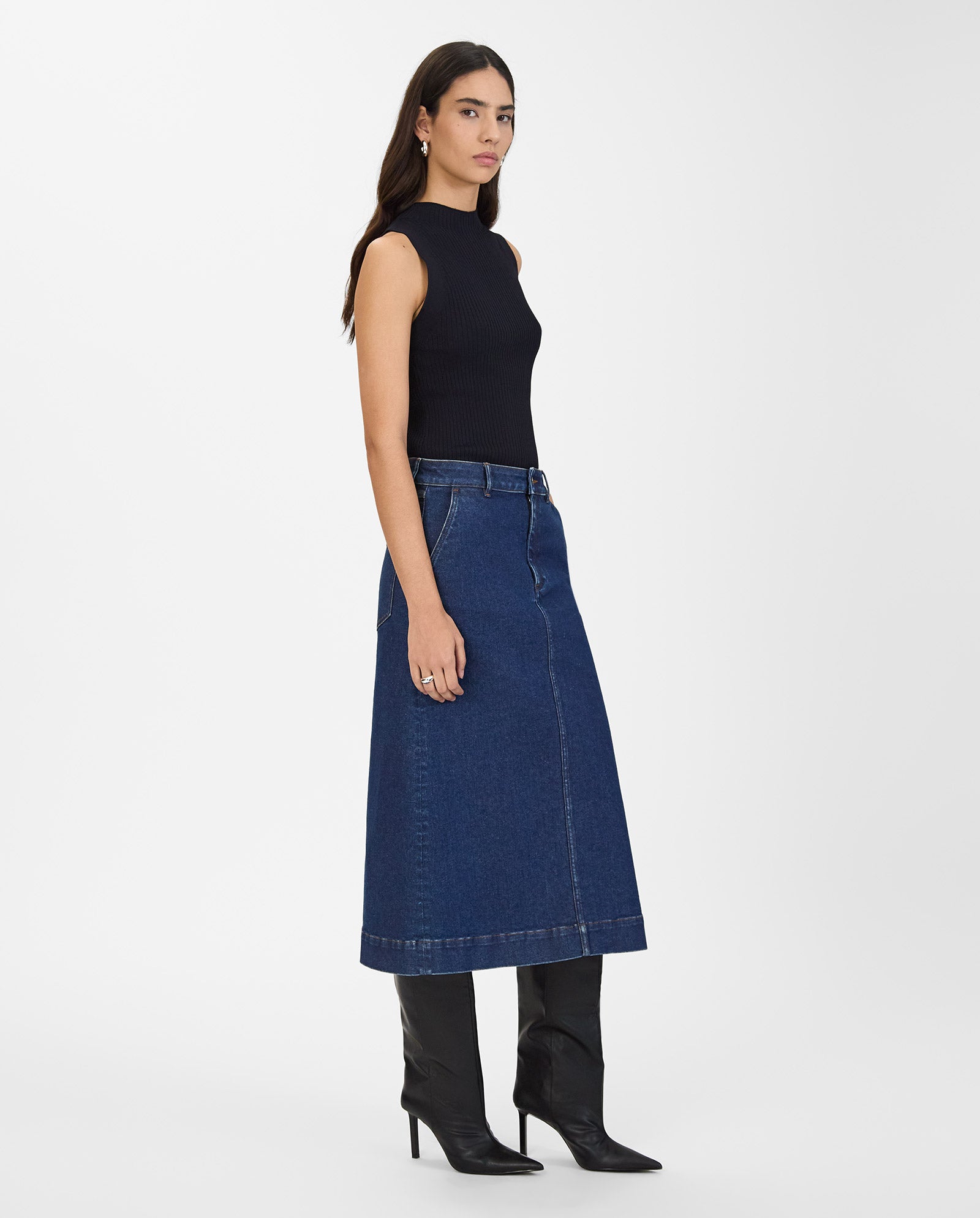 A woman wearing the IVY OAK STEFANIE Skirt—a long denim skirt with contrast stitching—paired with a sleeveless black top and black heeled boots, stands against a plain background.