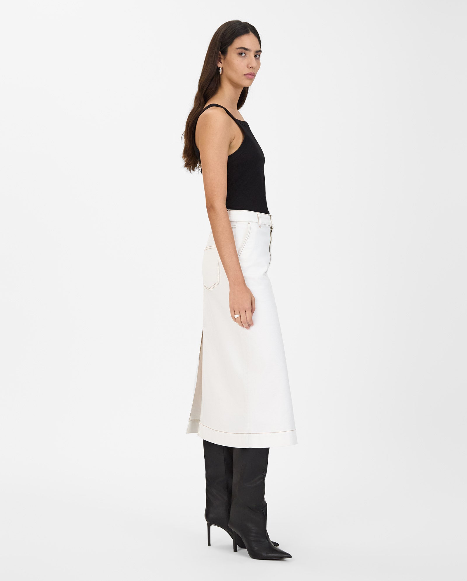 A woman stands in profile against a white background, wearing the IVY OAK STEFANIE Skirt—white denim with contrast stitching—paired with a black tank top and black heeled boots.