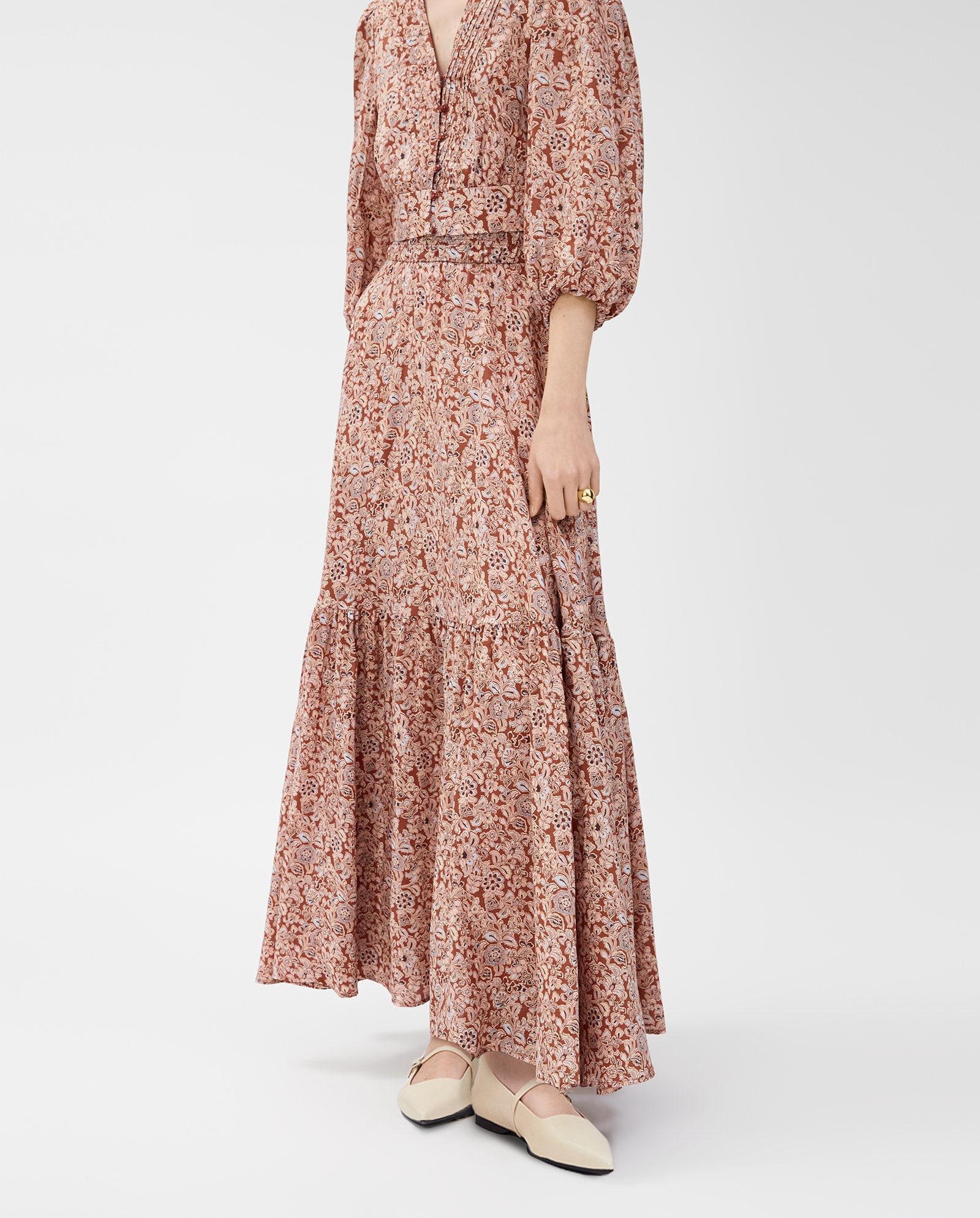 A woman wears the SANJA Skirt by IVY OAK— a long, flowy pink floral maxi with an elastic waistband— paired with cream-colored flat shoes.