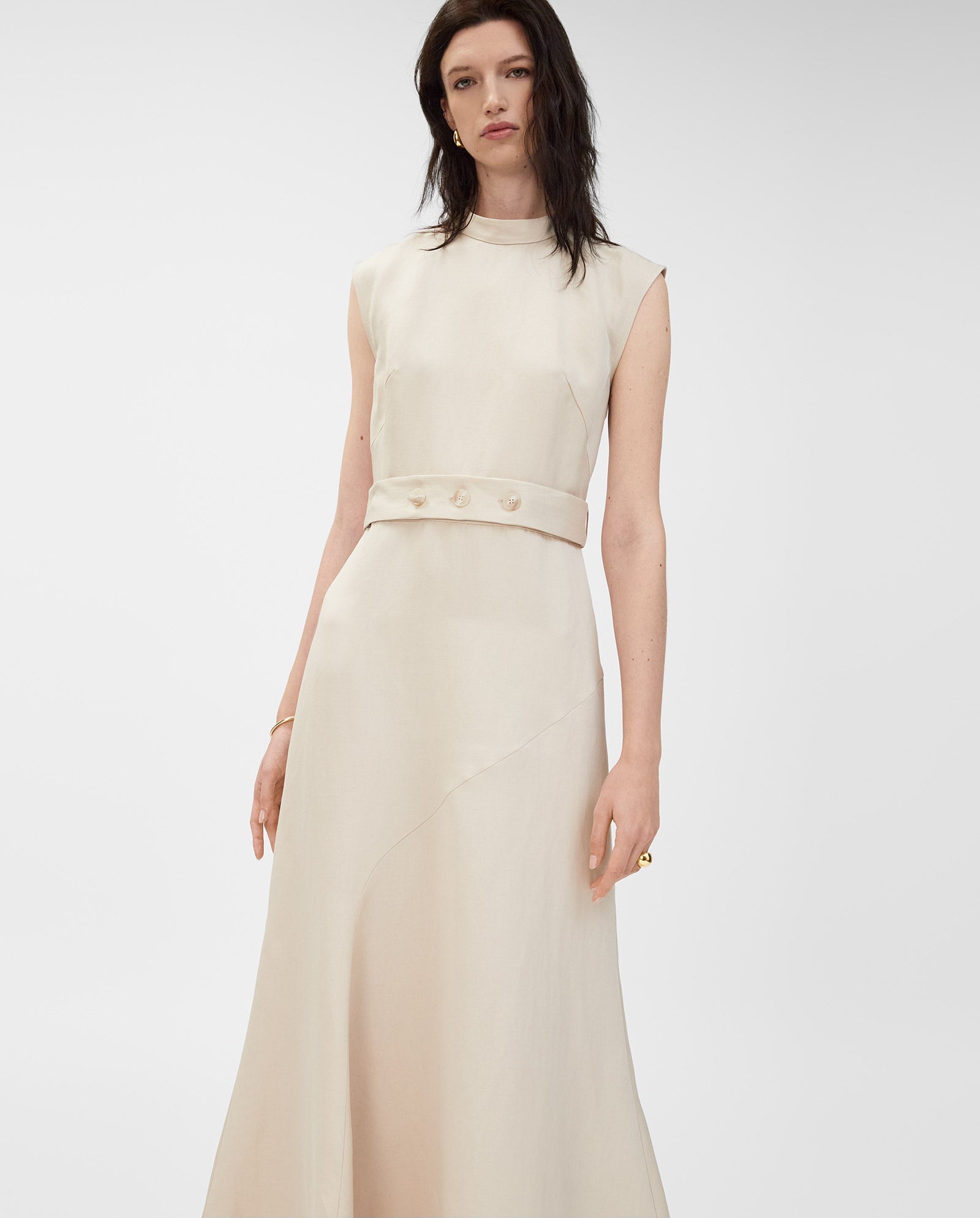 A woman wears the IVY OAK MANILA Dress, a sleeveless cream linen-blend piece with a belted waist, her soft wavy hair styled simply against a plain background.