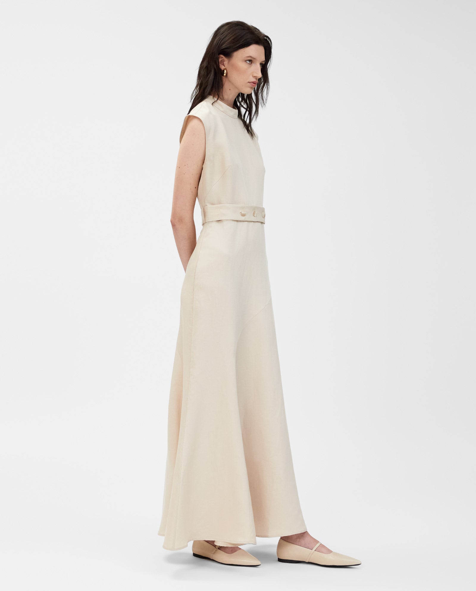 Woman wears the IVY OAK MANILA Dress, a sleeveless maxi in linen blend with belted waist and matching flats, posed against a white background. This chic dress brings effortless elegance to any occasion.