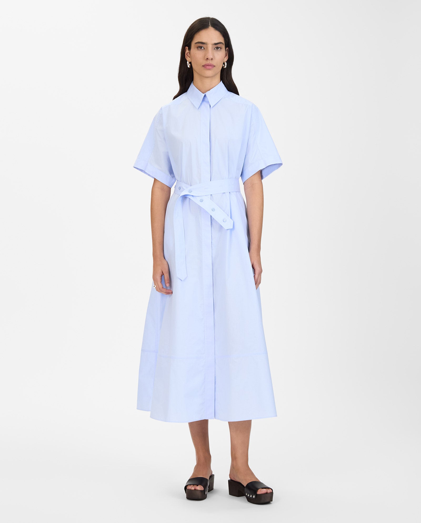 A woman wears the NORENA Dress by IVY OAK in light blue with short sleeves and black sandals, standing against a white background.