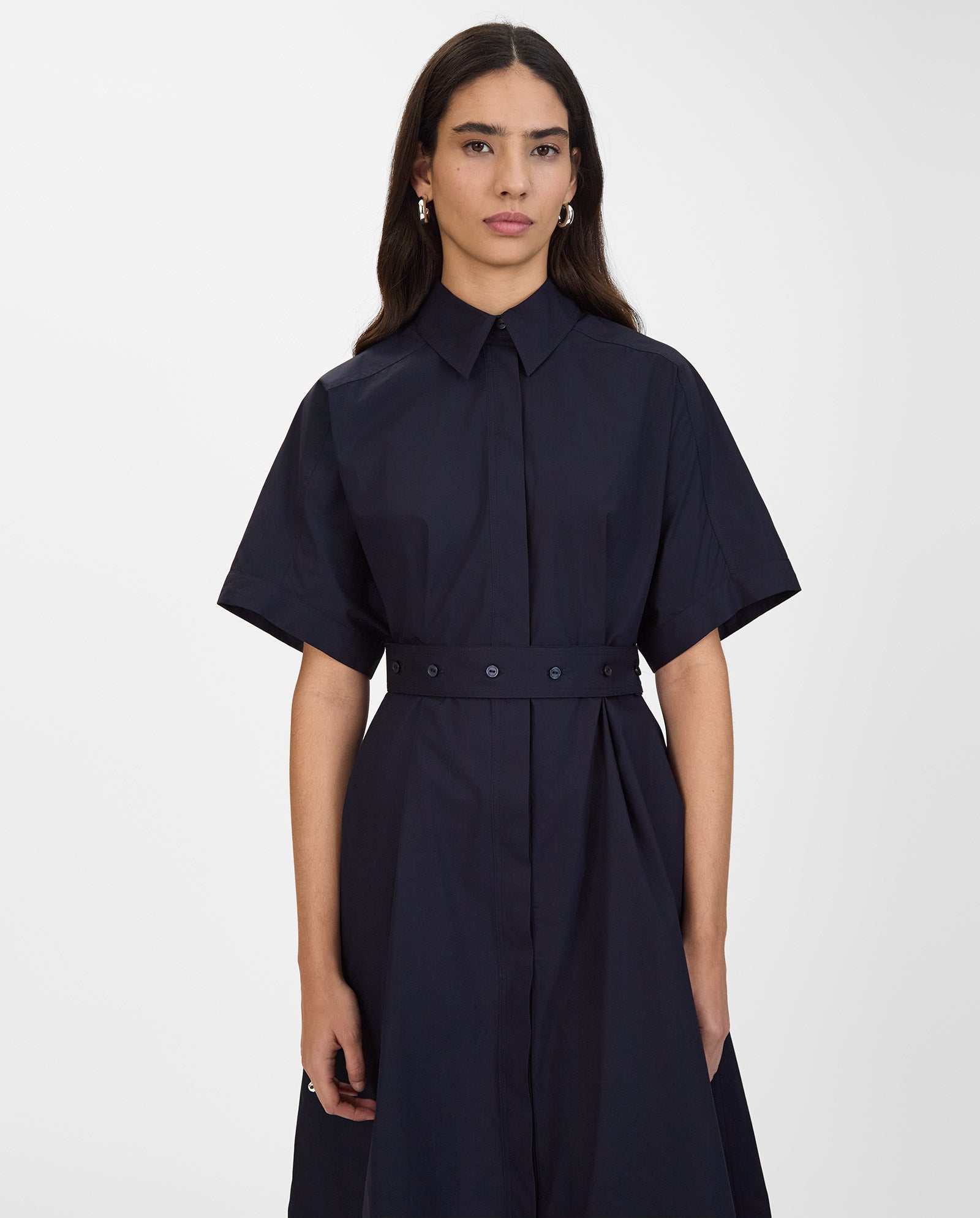 A woman wears the IVY OAK NORENA Dress, a dark navy shirt dress with short sleeves and a belt, standing against a plain white background.