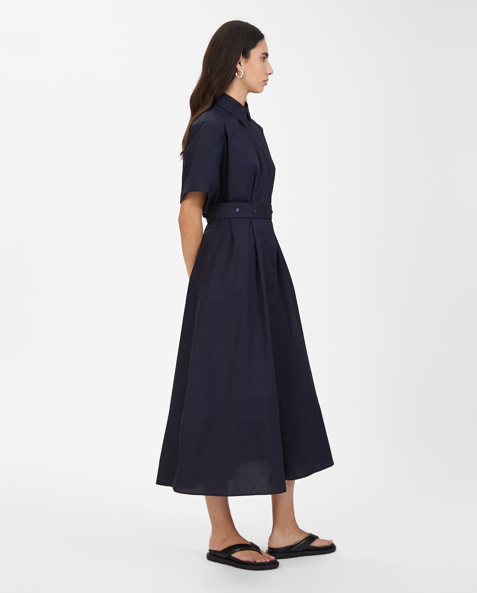 A woman stands in profile against a white background, wearing the IVY OAK NORENA Dress in navy with short sleeves and a belted waist, paired with black sandals.