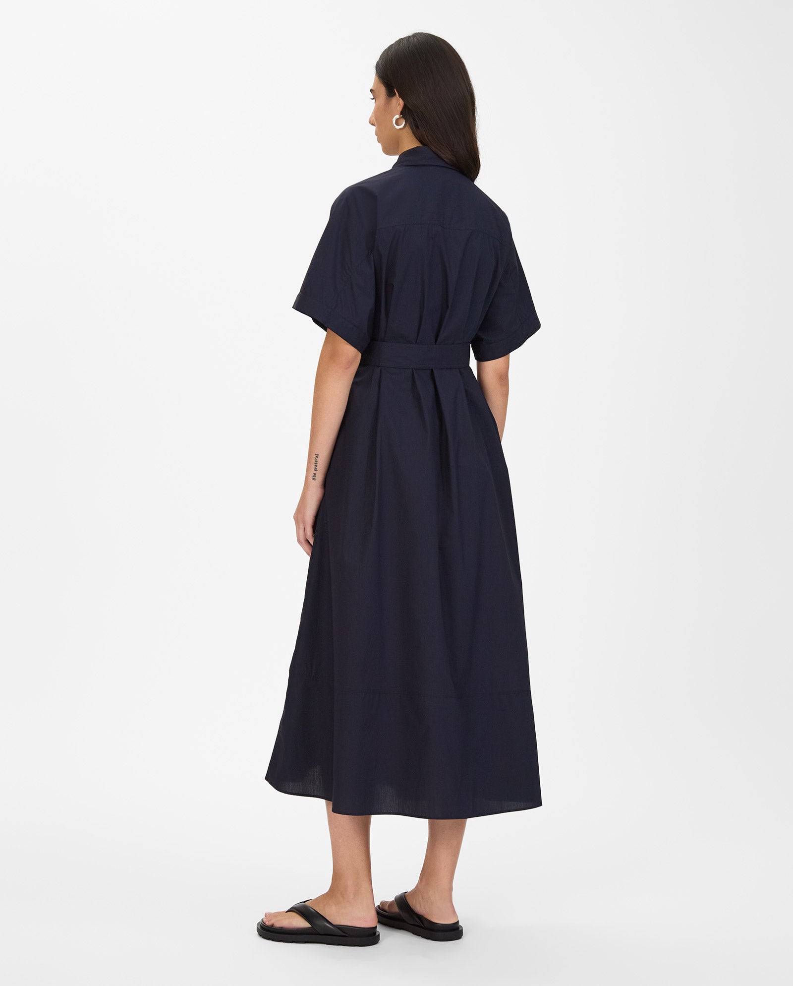 Woman wearing the IVY OAK NORENA Dress in navy blue, featuring short sleeves, a belt, and midi length, paired with black sandals and facing away.