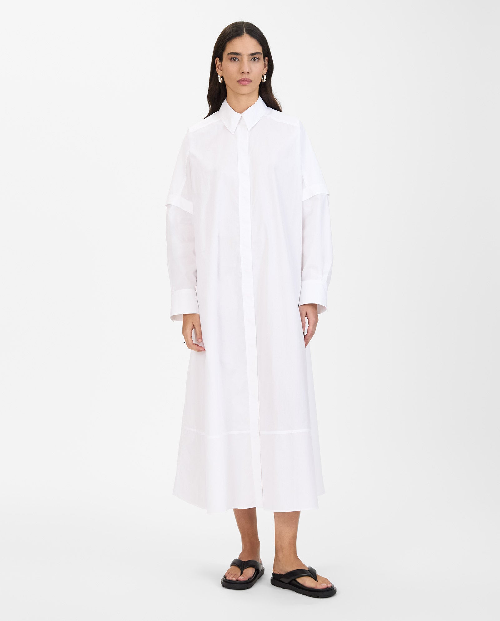 A woman wears the NORENA Dress by IVY OAK—a long white shirt dress with detachable sleeves—paired with black sandals, standing against a plain background.