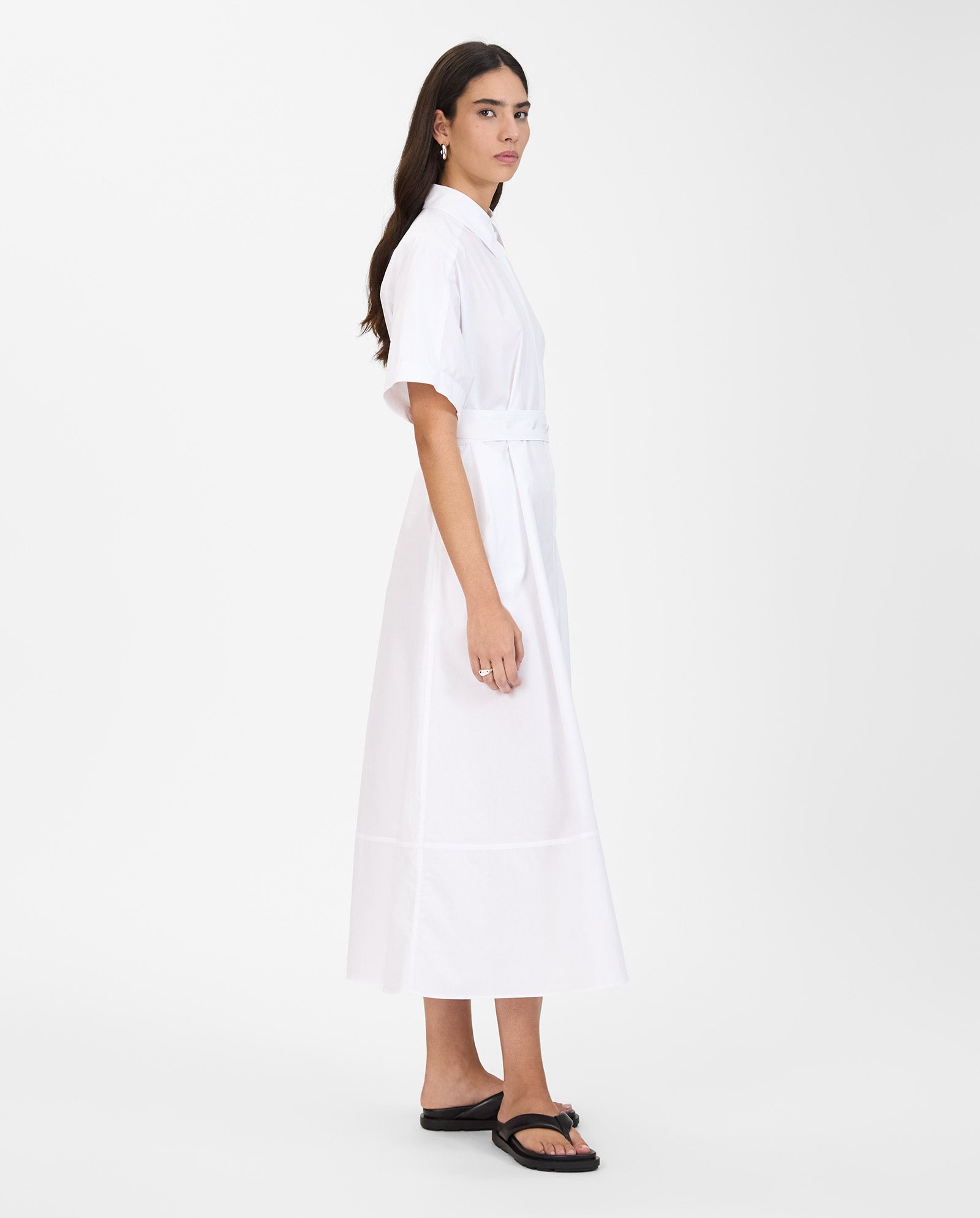 A woman stands sideways against a white background wearing the IVY OAK NORENA Dress, a white shirt dress with detachable sleeves, paired with black sandals.