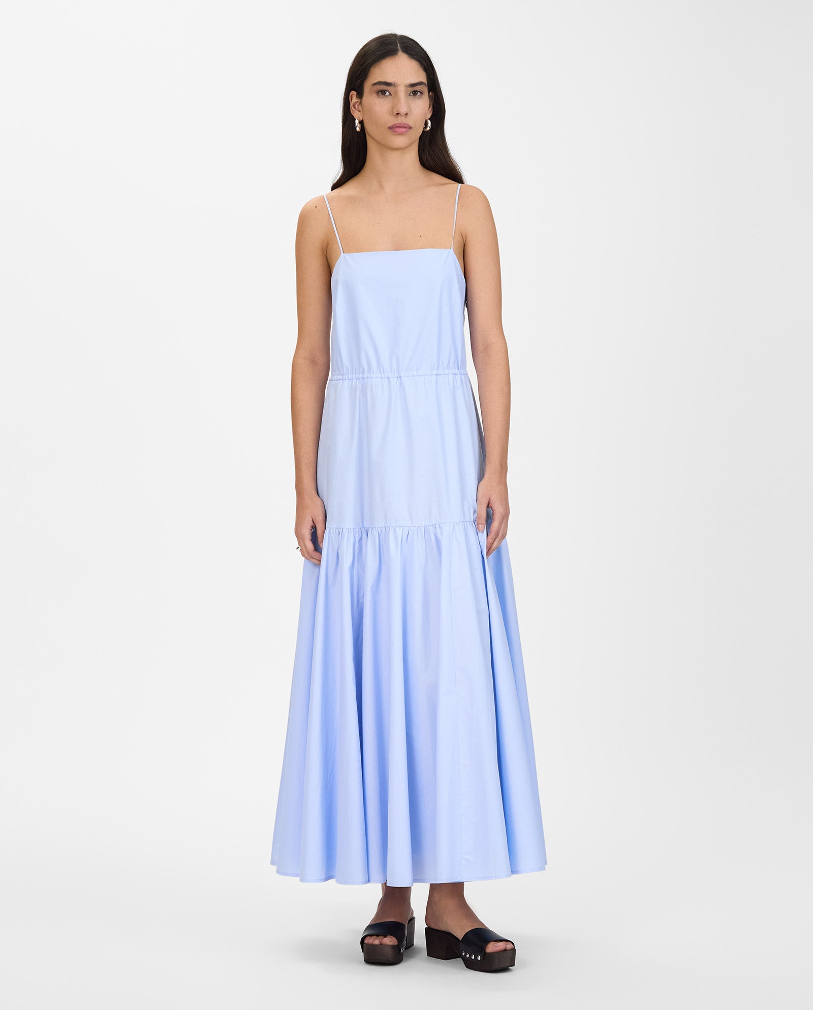 Woman wears the IVY OAK NICOLINA Dress, a light blue sleeveless maxi with adjustable straps, standing against a plain white background.