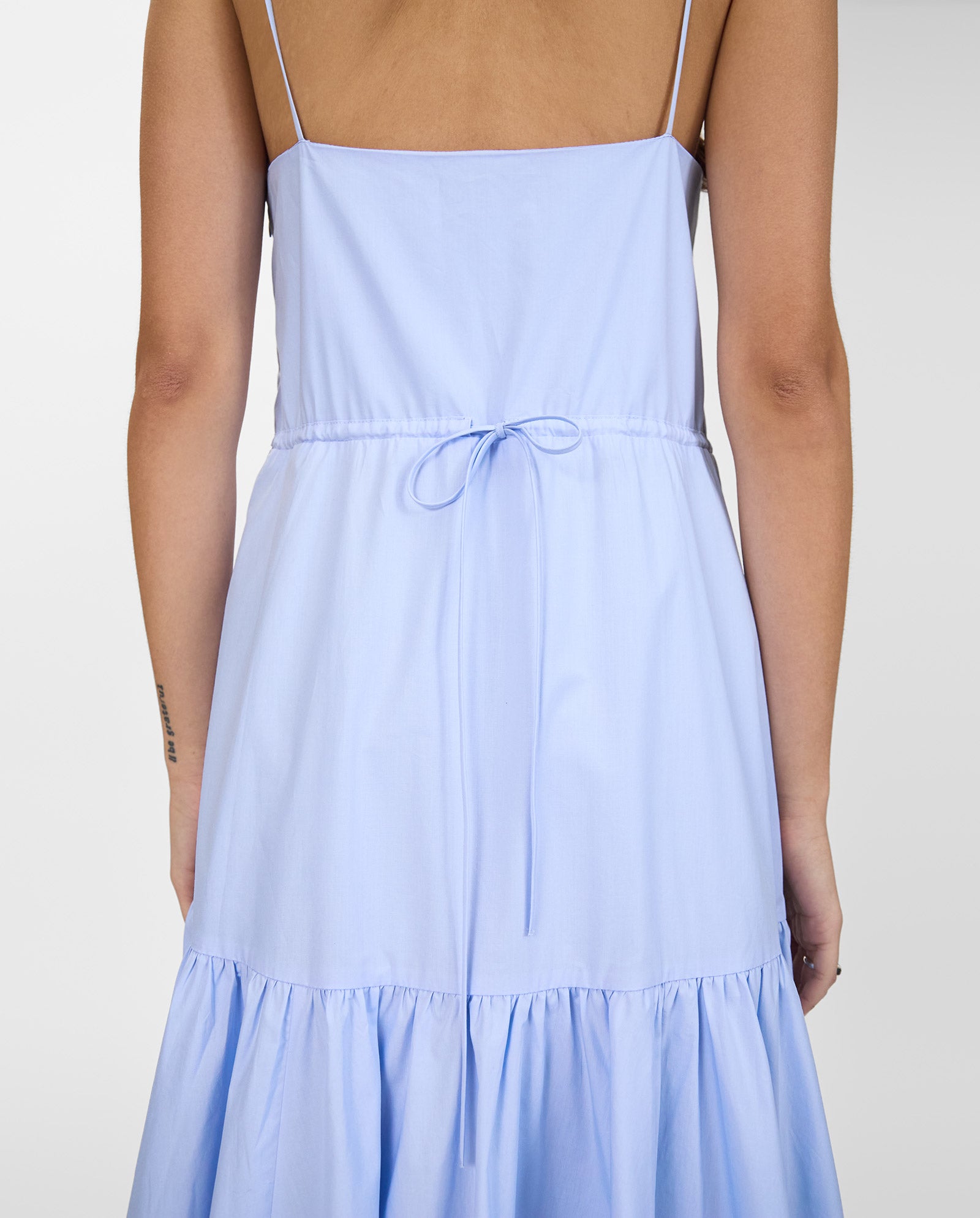 Woman wearing the IVY OAK NICOLINA Dress, a light blue sleeveless style with adjustable straps and a tie waist, shown from the back.