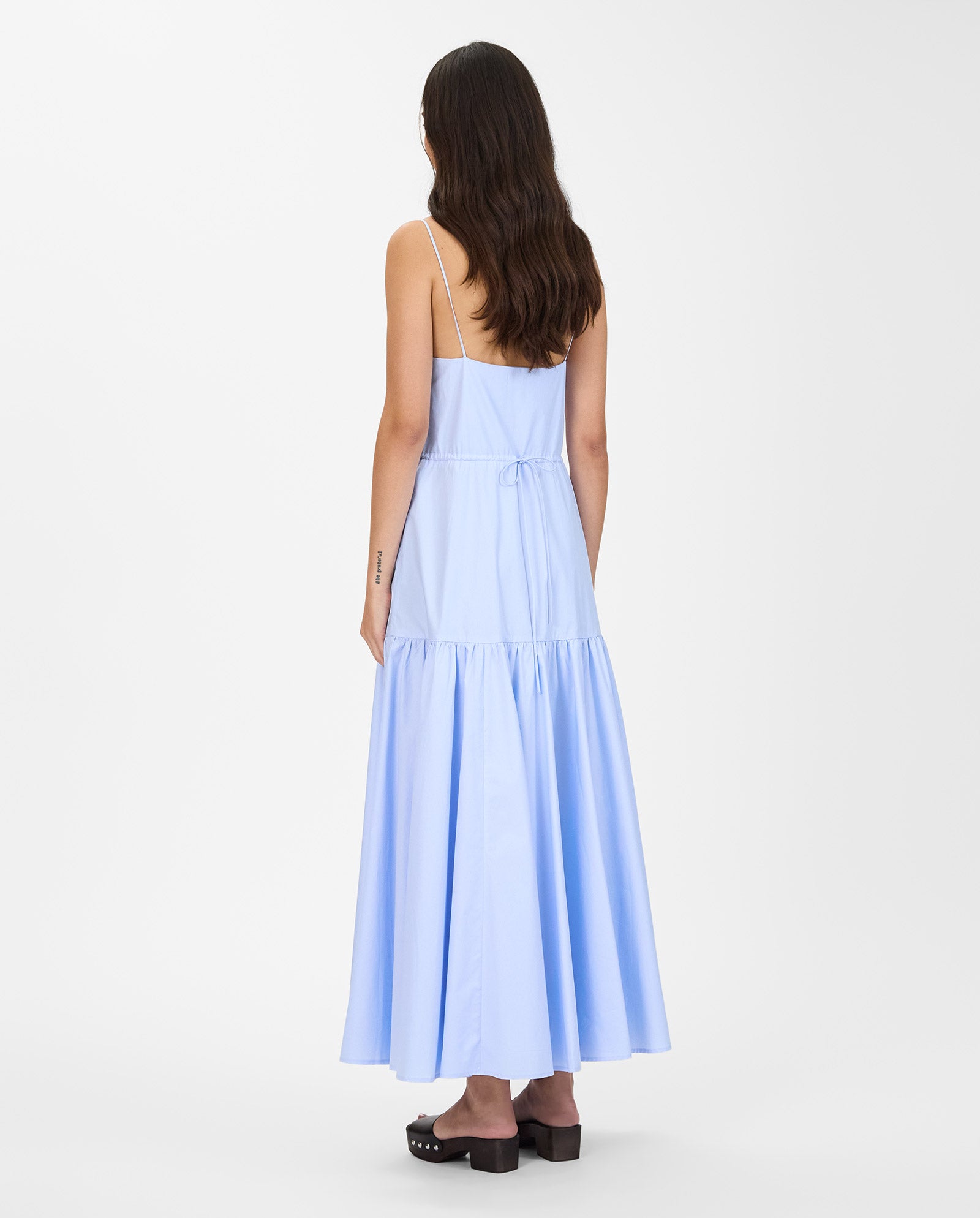 A woman, seen from behind, wears the IVY OAK NICOLINA Dress in light blue with adjustable straps and a maxi length, paired with black clogs.