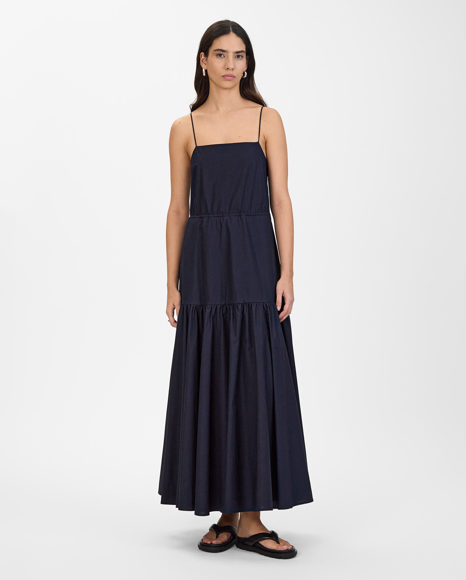 A woman wears the IVY OAK NICOLINA Dress, a navy blue maxi with adjustable straps, and black sandals, standing against a white background.
