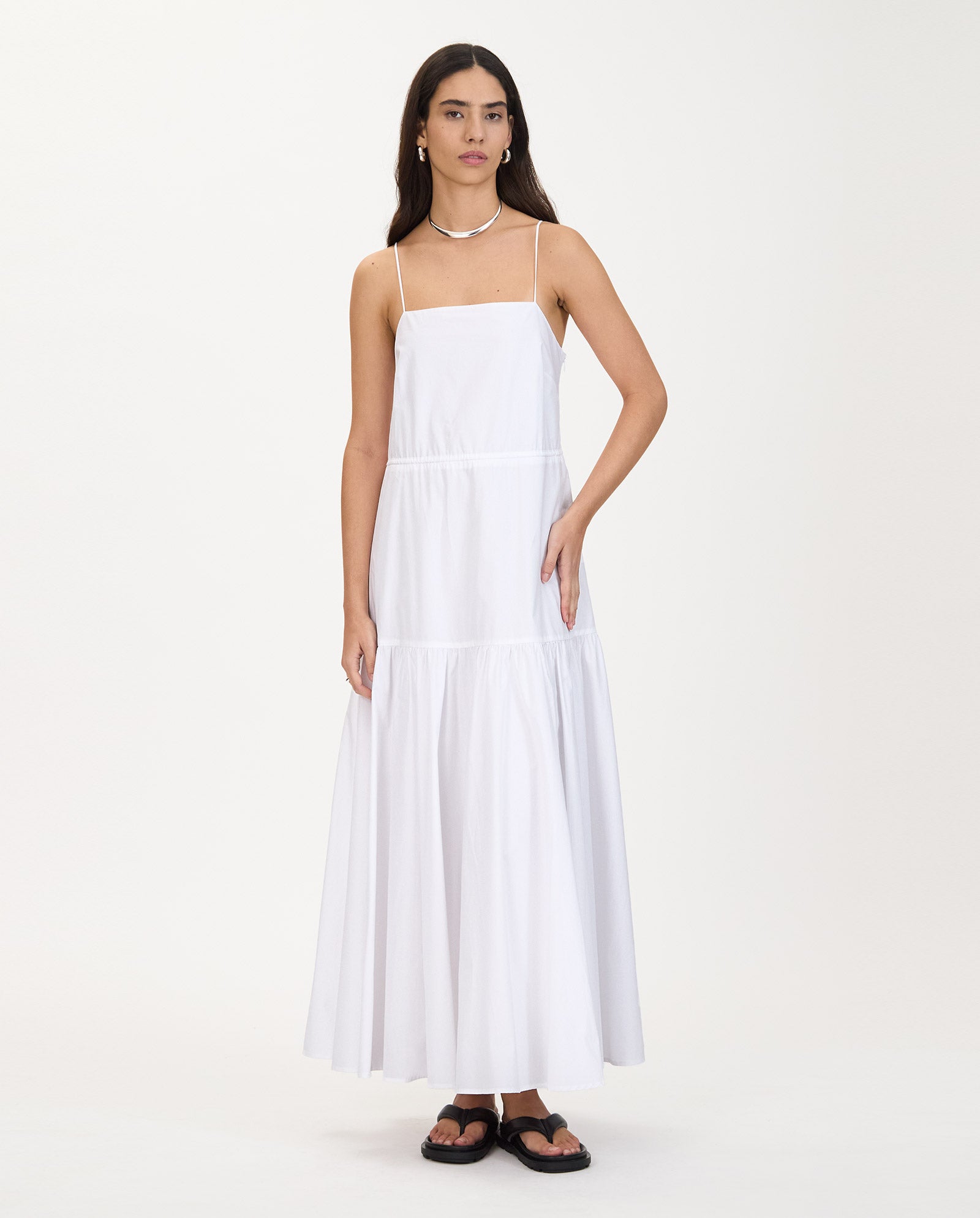 Woman in a long, white IVY OAK NICOLINA Dress with maxi length, A-line silhouette, and thin straps, styled with sandals and a necklace, standing on white.