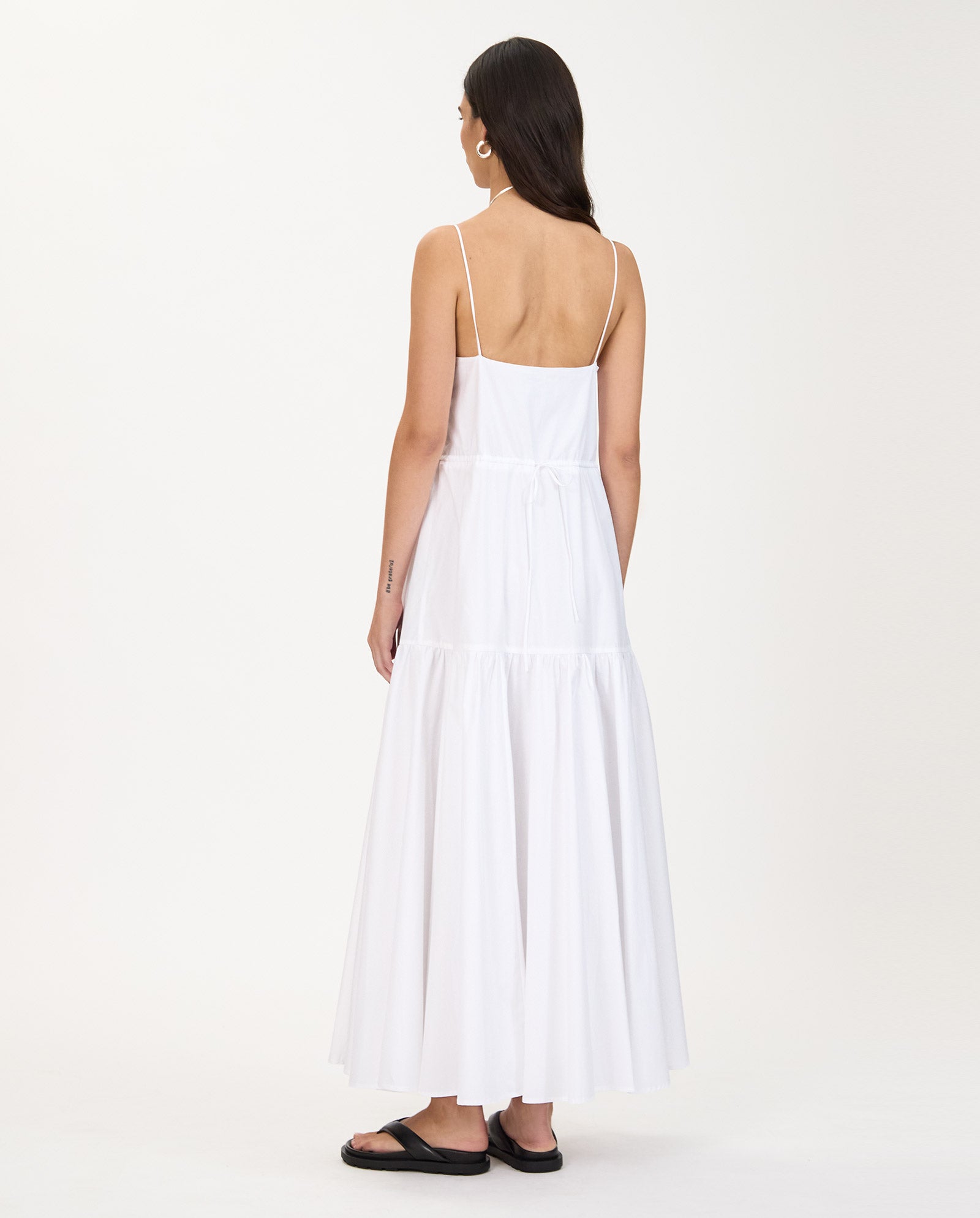 Woman in a long white sleeveless IVY OAK NICOLINA Dress with an A-line silhouette and maxi length, wearing black sandals, standing with her back to the camera.