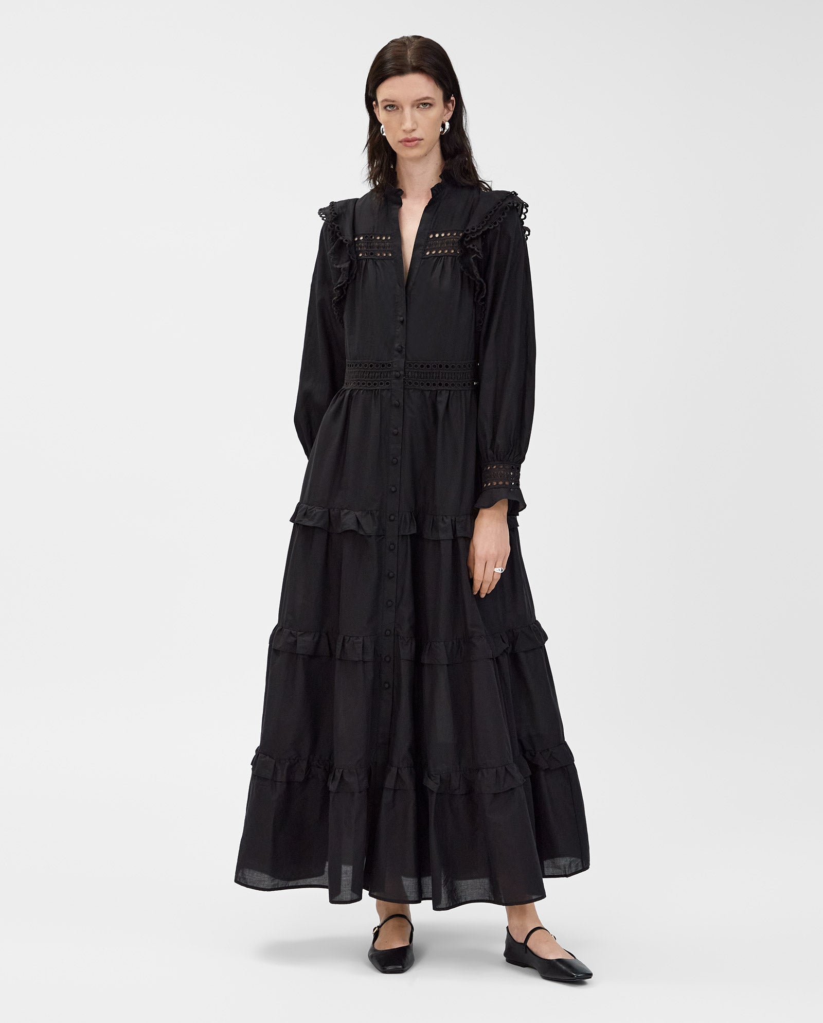 A woman wears the IVY OAK DENISA Dress, a long black cotton voile design with lace tape, ruffles, buttoned front, and lace details. She completes her look with classic black flats.