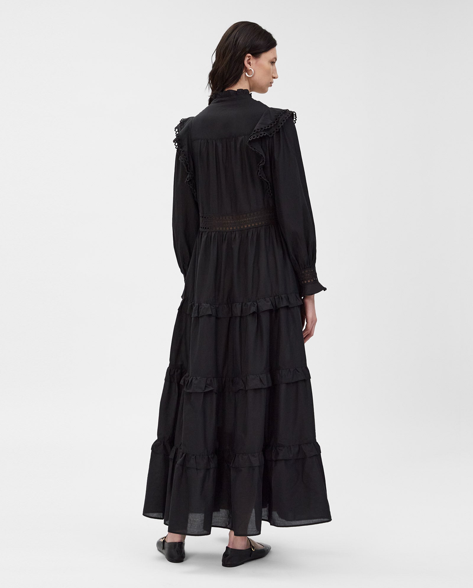 A woman stands facing away from the camera in the long black DENISA Dress by IVY OAK, featuring ruffled details and lace tape accents, crafted from cotton voile.