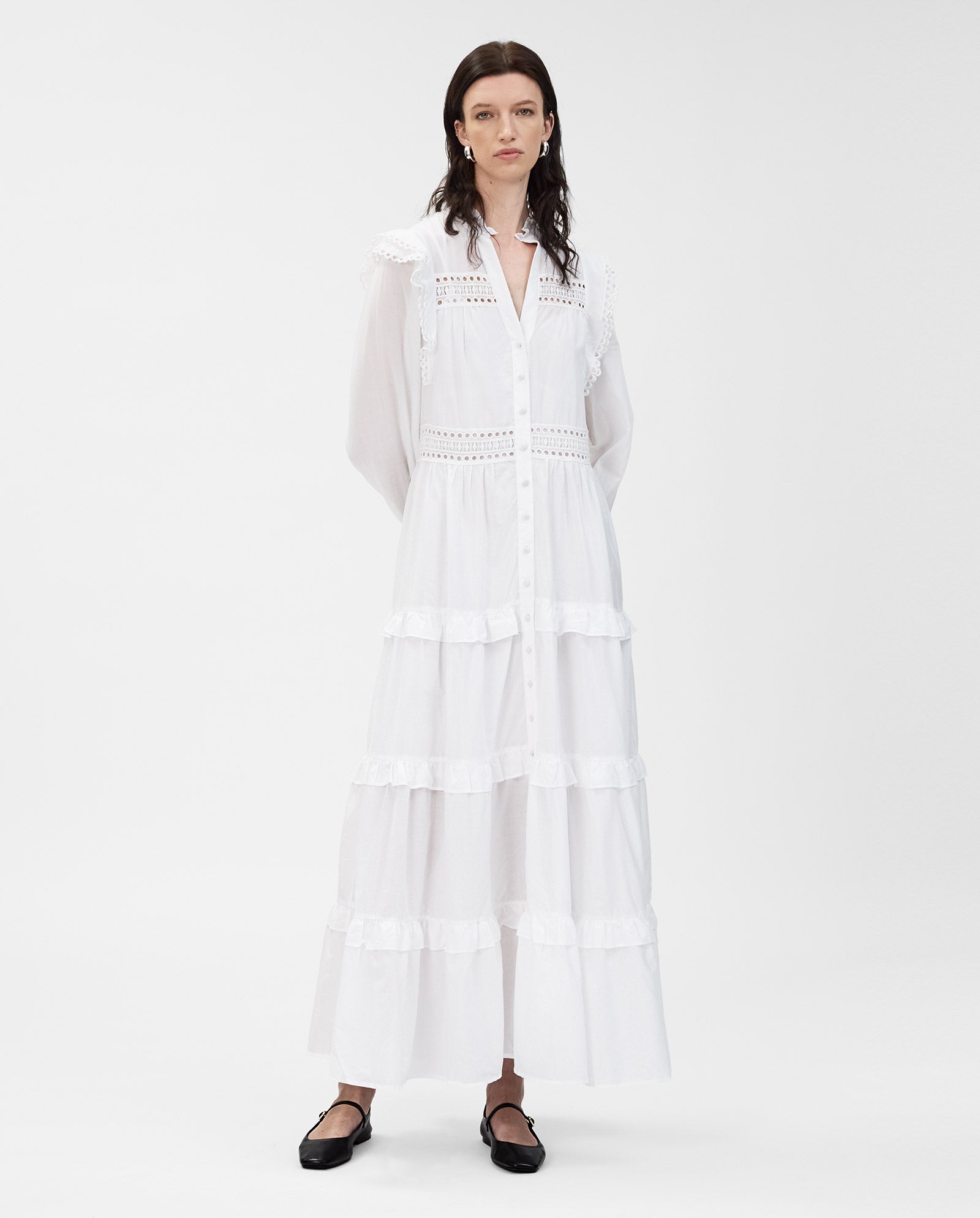 A woman wears the IVY OAK DENISA Dress—a long white cotton voile dress with ruffles, lace tape details, and fabric-covered buttons—paired with black flats, standing against a white background.