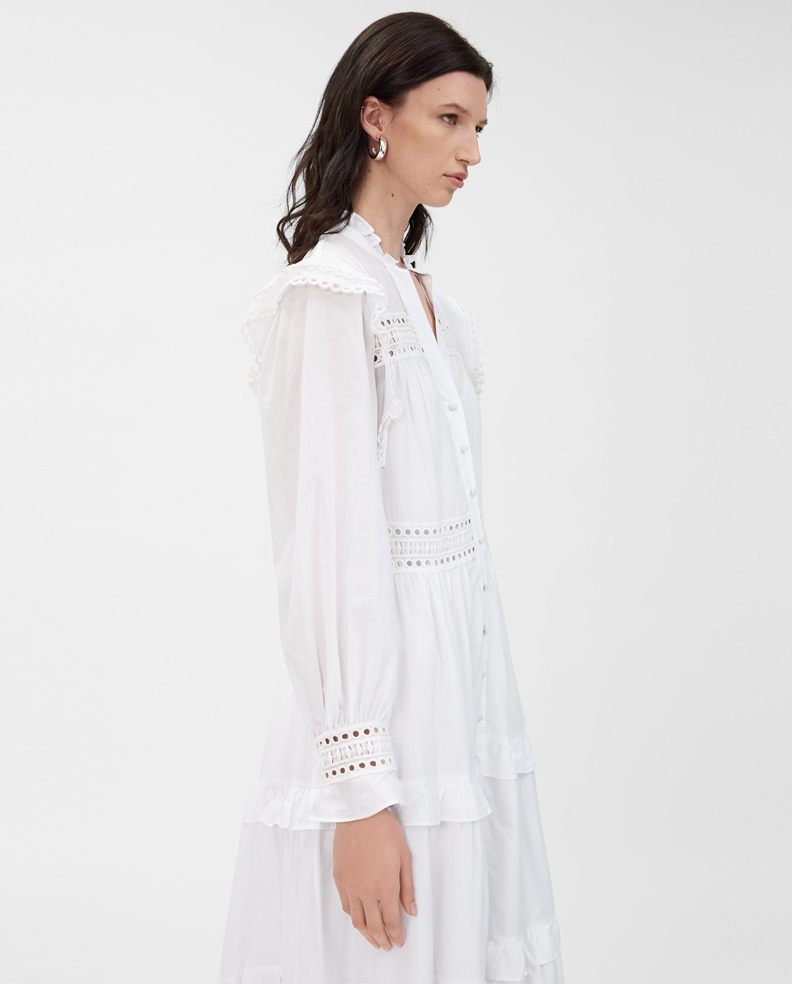 A woman in the IVY OAK DENISA Dress, made of white cotton voile with fabric-covered buttons and lace tape accents, stands in profile against a plain background.