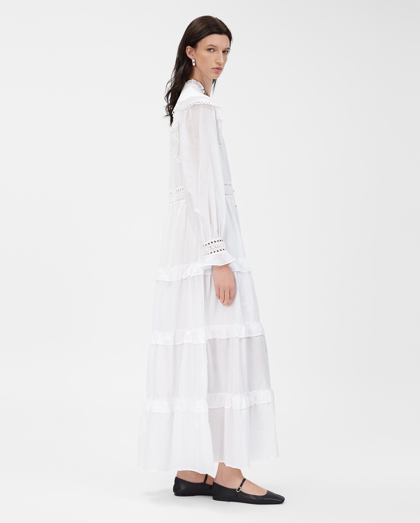 A woman in the IVY OAK DENISA dress—a long, white cotton voile design with lace tape details and fabric-covered buttons—wears black flats and stands against a white background.