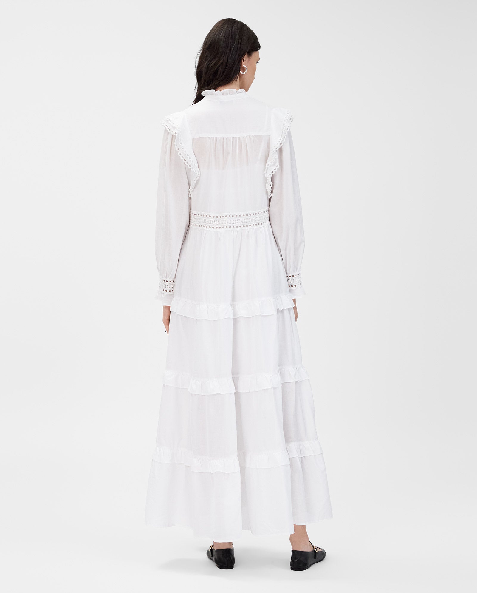 A woman stands with her back to the camera, wearing the IVY OAK DENISA Dress in long white cotton voile with fabric-covered buttons and black shoes.