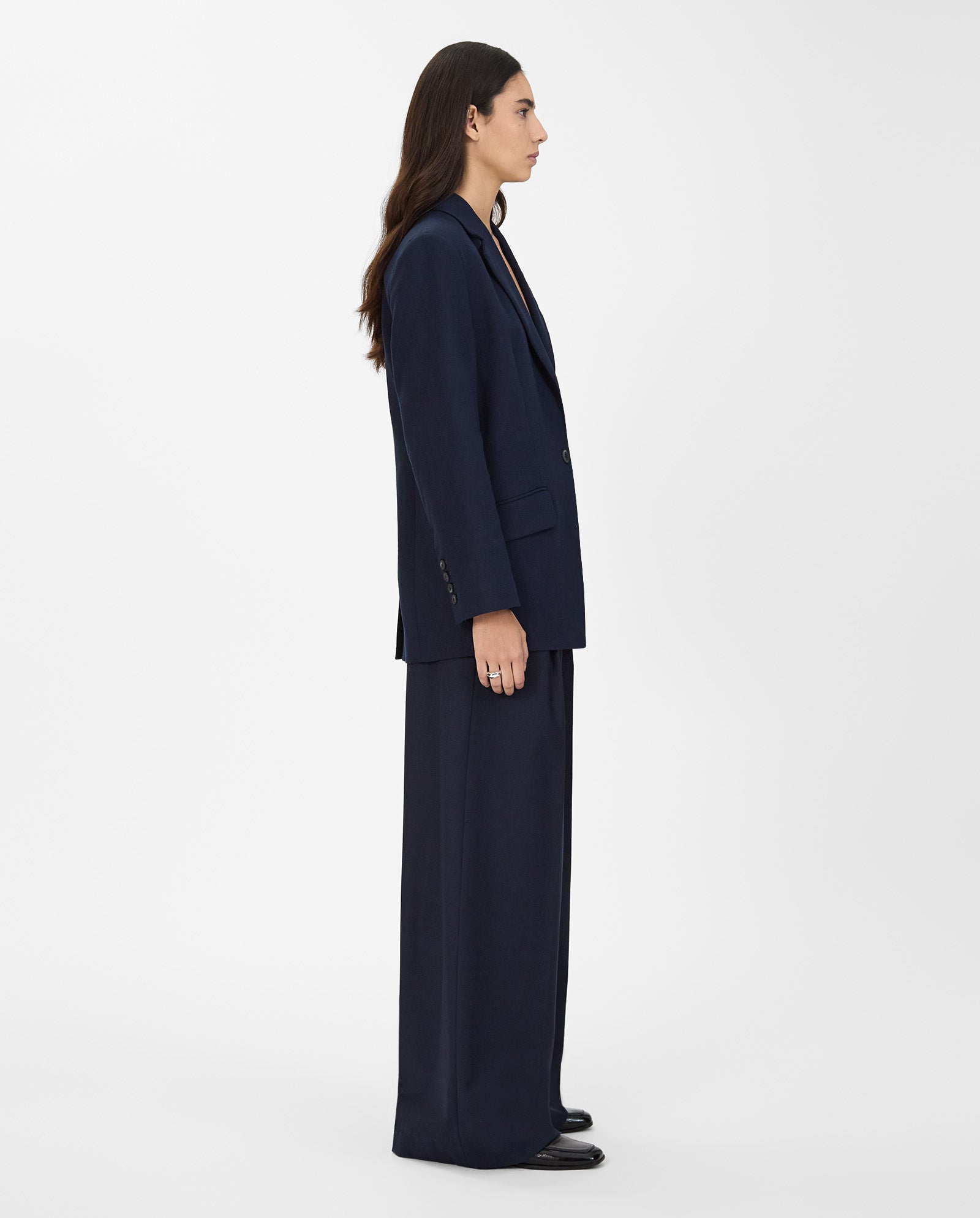 A person with long hair models the IVY OAK JUNA Blazer and matching trousers in navy, standing in profile against a plain white background.