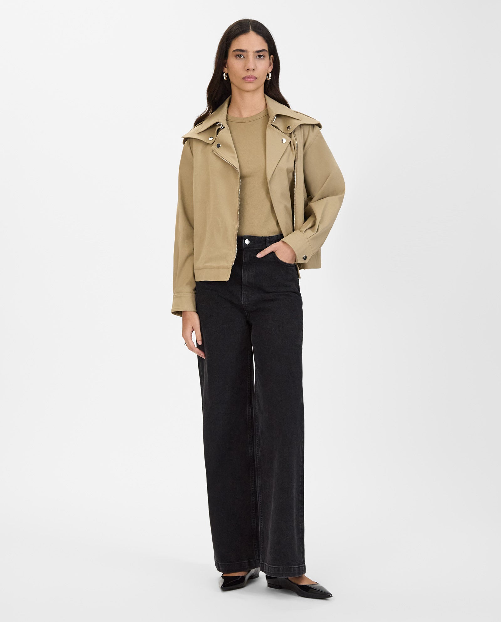 A woman wears the IVY OAK JOANNE Jacket, a tan trench with metal trimmings, paired with a beige top, black wide-leg jeans, and black shoes, standing against a white background.