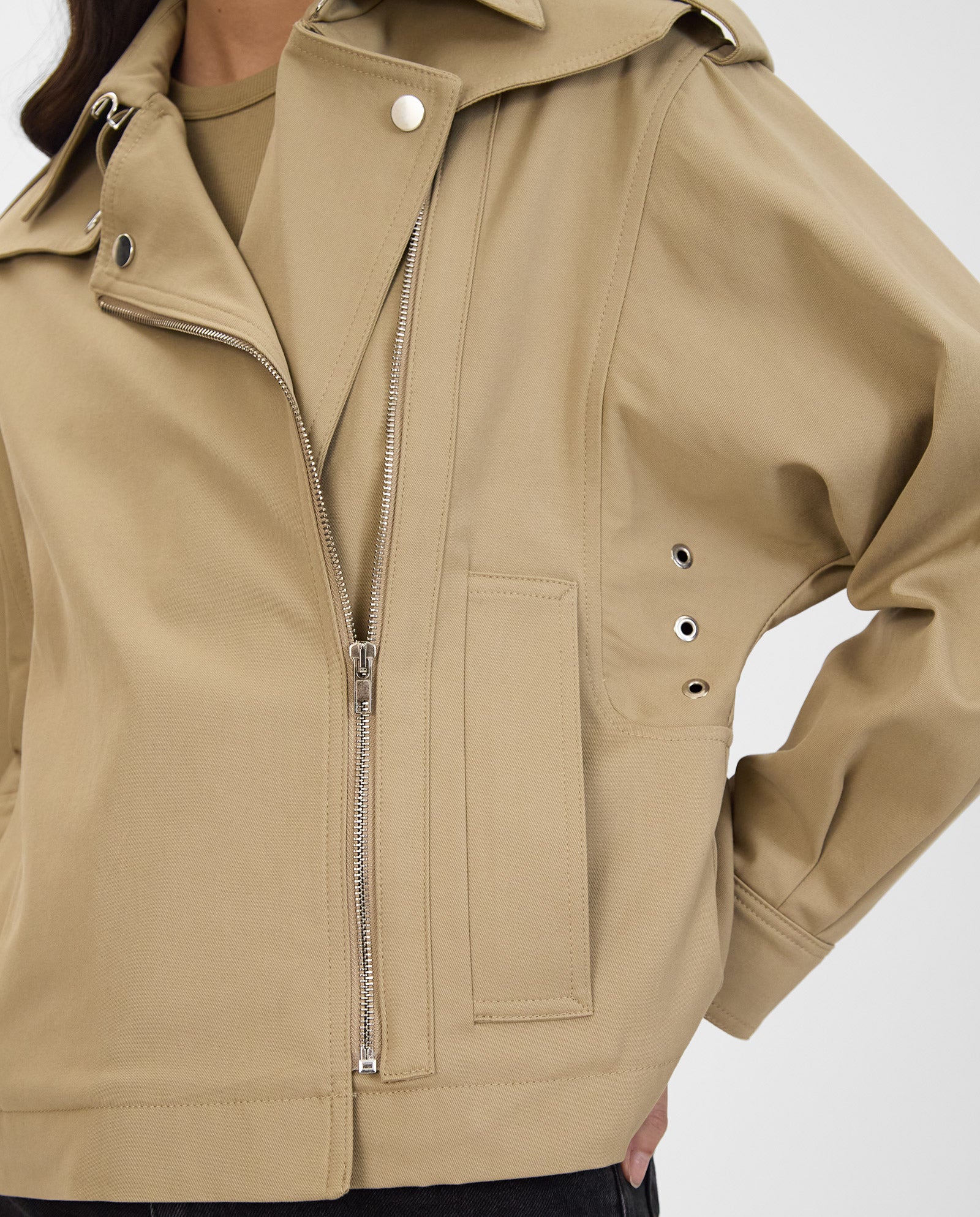 A person wearing the IVY OAK JOANNE Jacket, a tan trench with yoke flaps, asymmetrical zipper, and silver snap buttons, poses with one hand on hip.