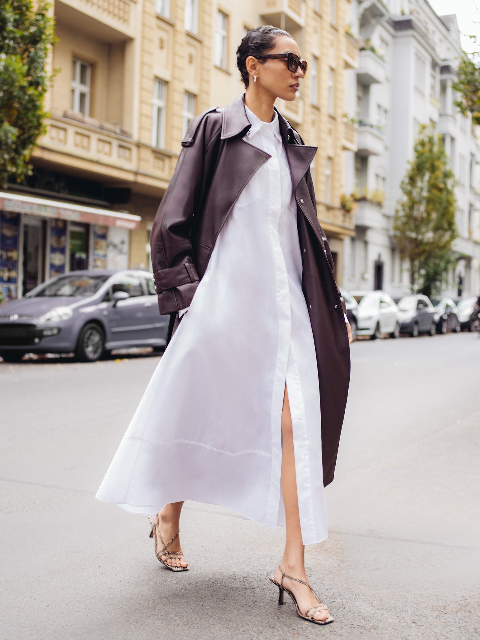 A woman wearing IVY OAKs Adelaine Sunglasses, crafted from biodegradable acetate, strolls a city street in a long white dress, dark trench coat, and heeled sandals.