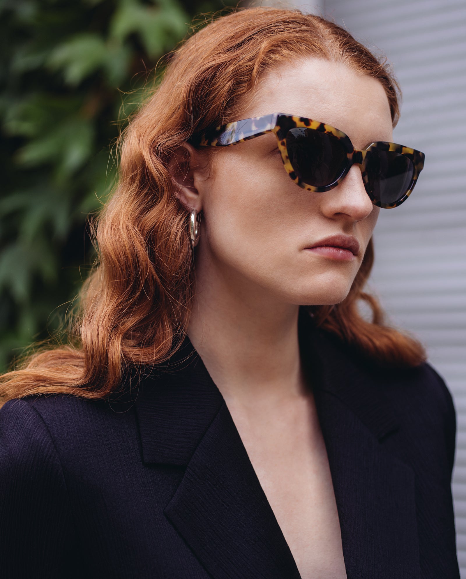Woman with wavy red hair wears IVY OAK Addison Sunglasses with tortoiseshell frames and UV protection, paired with hoop earrings and a black blazer outdoors.