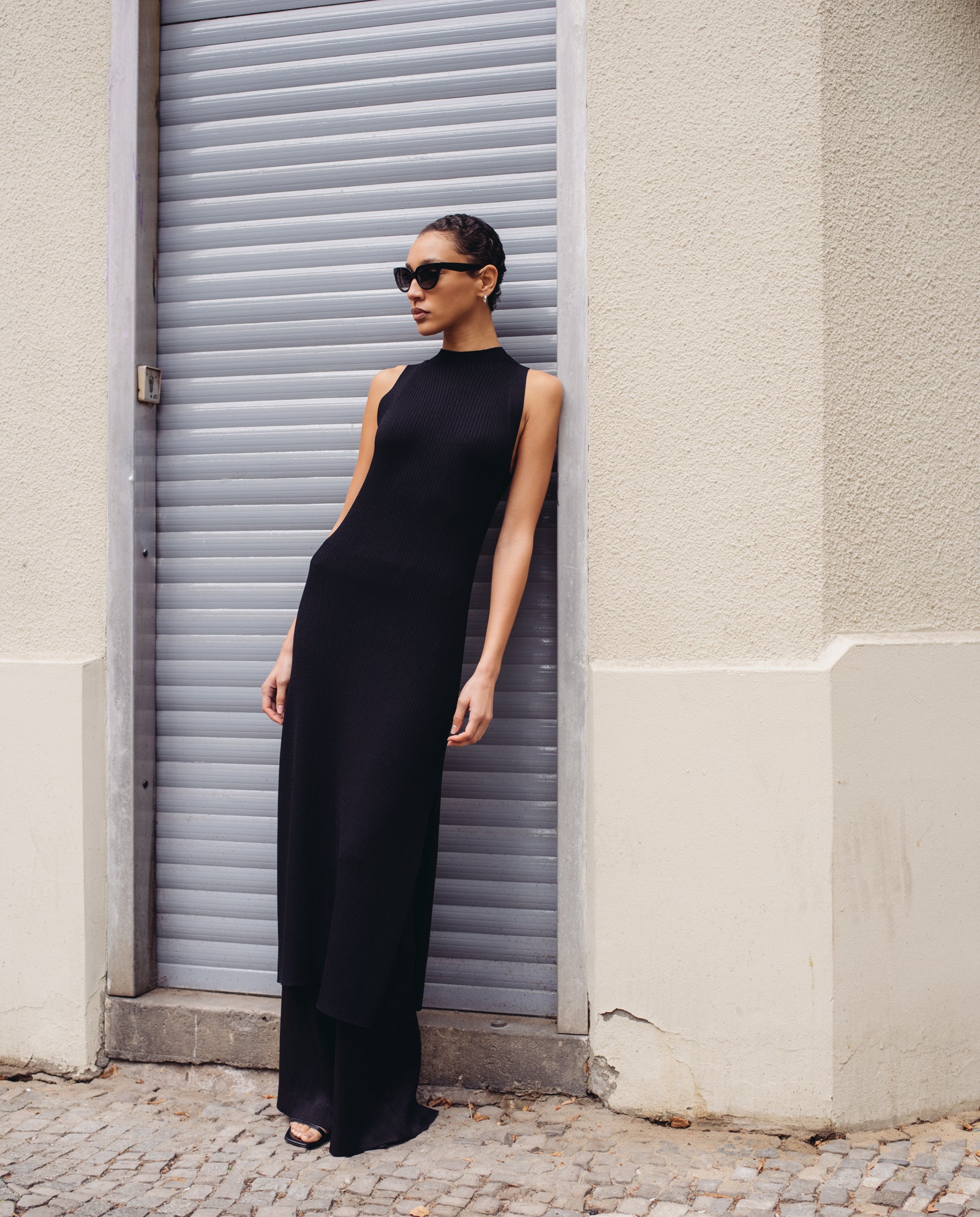 A woman wearing IVY OAK’s Addison Sunglasses and a black sleeveless dress leans against a gray metal shutter on a city street.