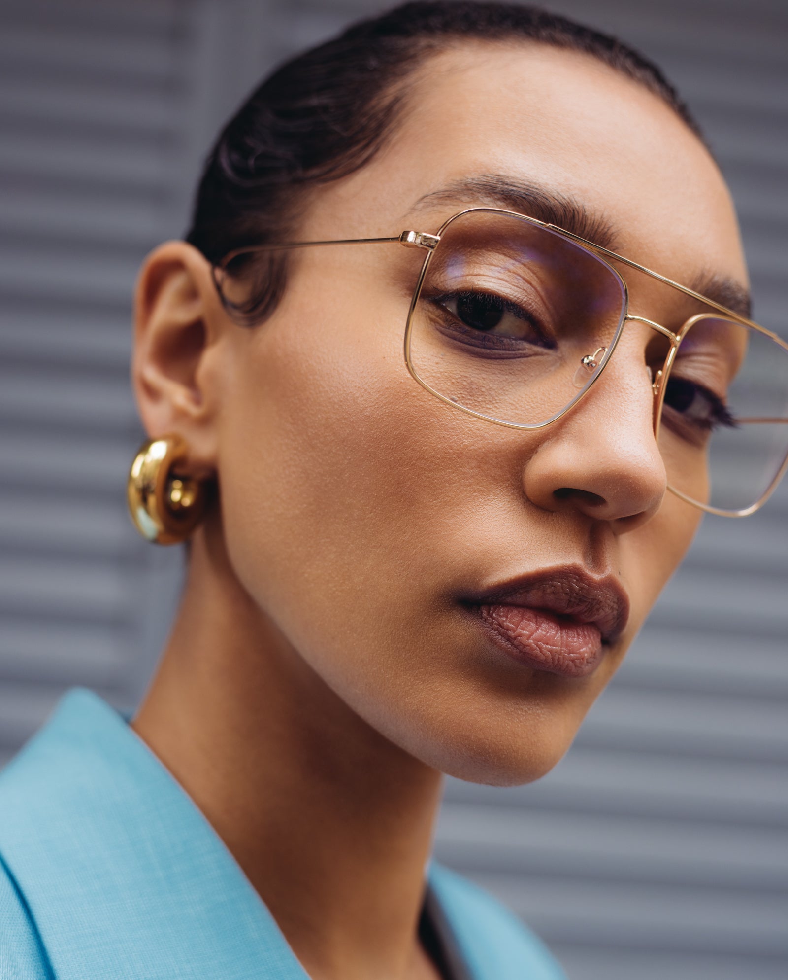 A woman wears IVY OAK’s Augusta Glasses with gold rims and blue light filter, paired with gold hoop earrings and a light blue blazer, gazing ahead with confidence.