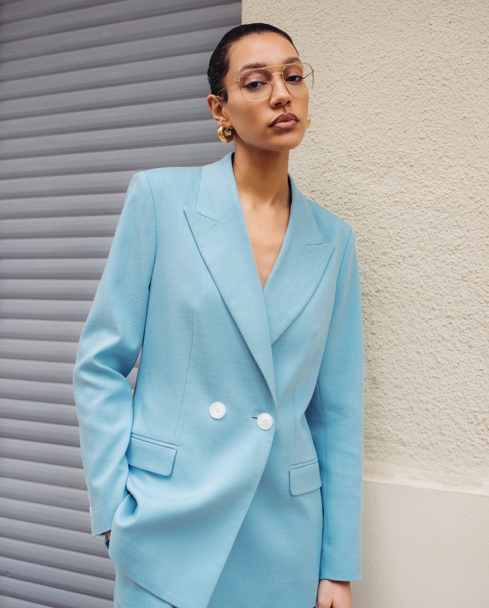 A woman in a light blue suit jacket and gold hoop earrings wears IVY OAKs latest Augusta Glasses, standing against a textured wall and shutter.