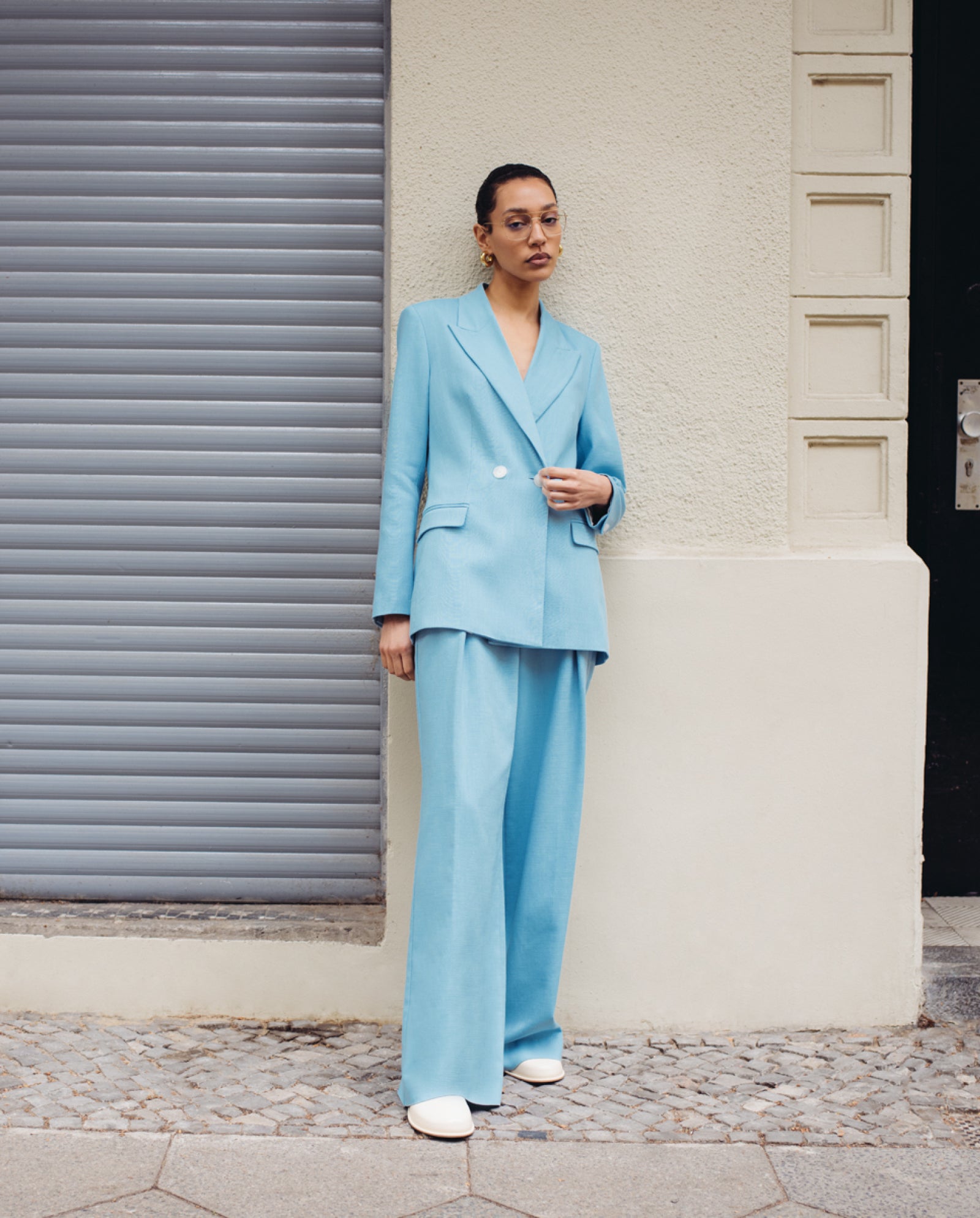 A woman in a light blue pantsuit and white shoes stands against a cream wall and gray shutter, modeling IVY OAK’s Augusta Glasses—an elegant eyewear collection crafted from recycled materials.
