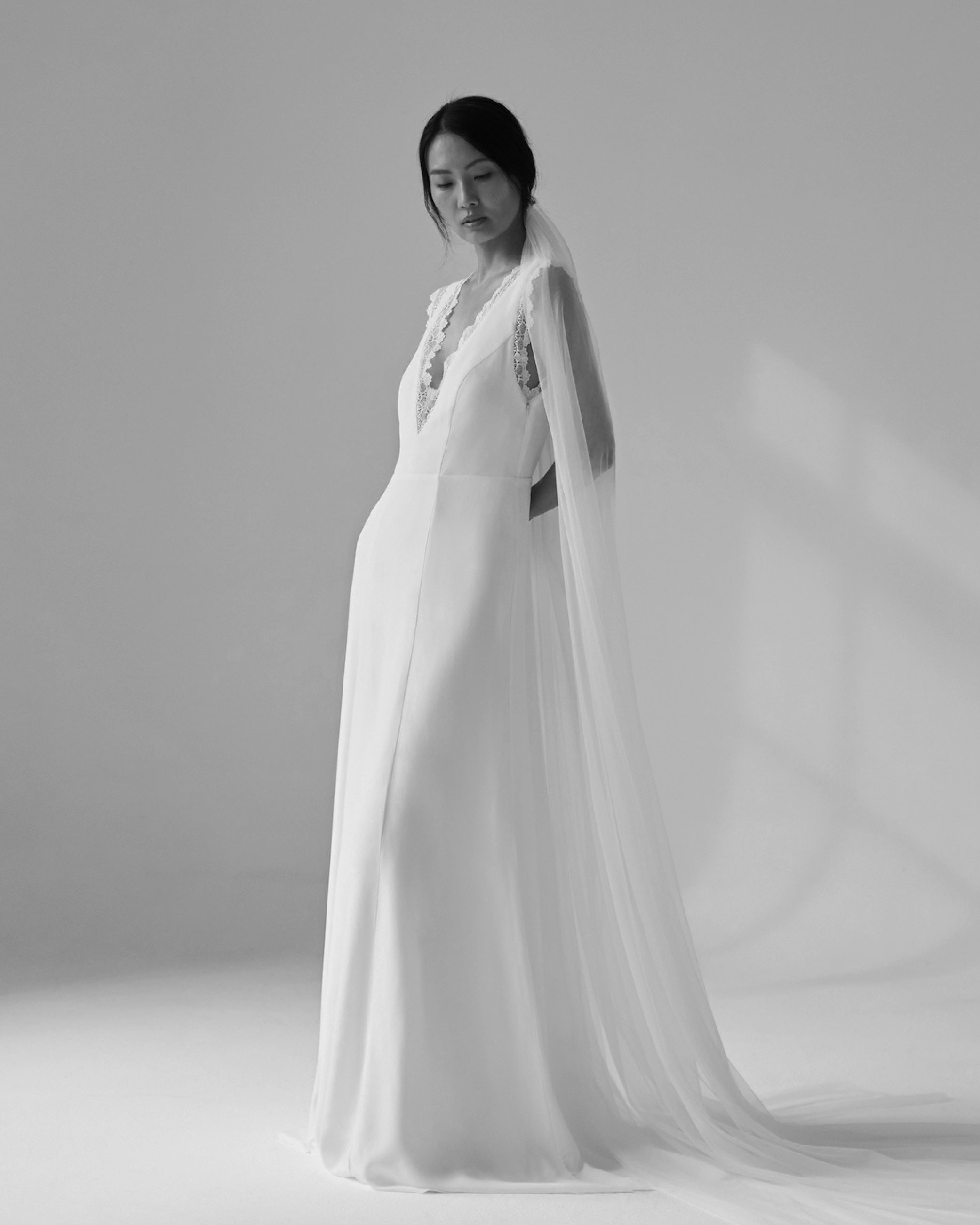 Woman in a long, elegant white bridal gown with a veil, standing in soft, natural light.