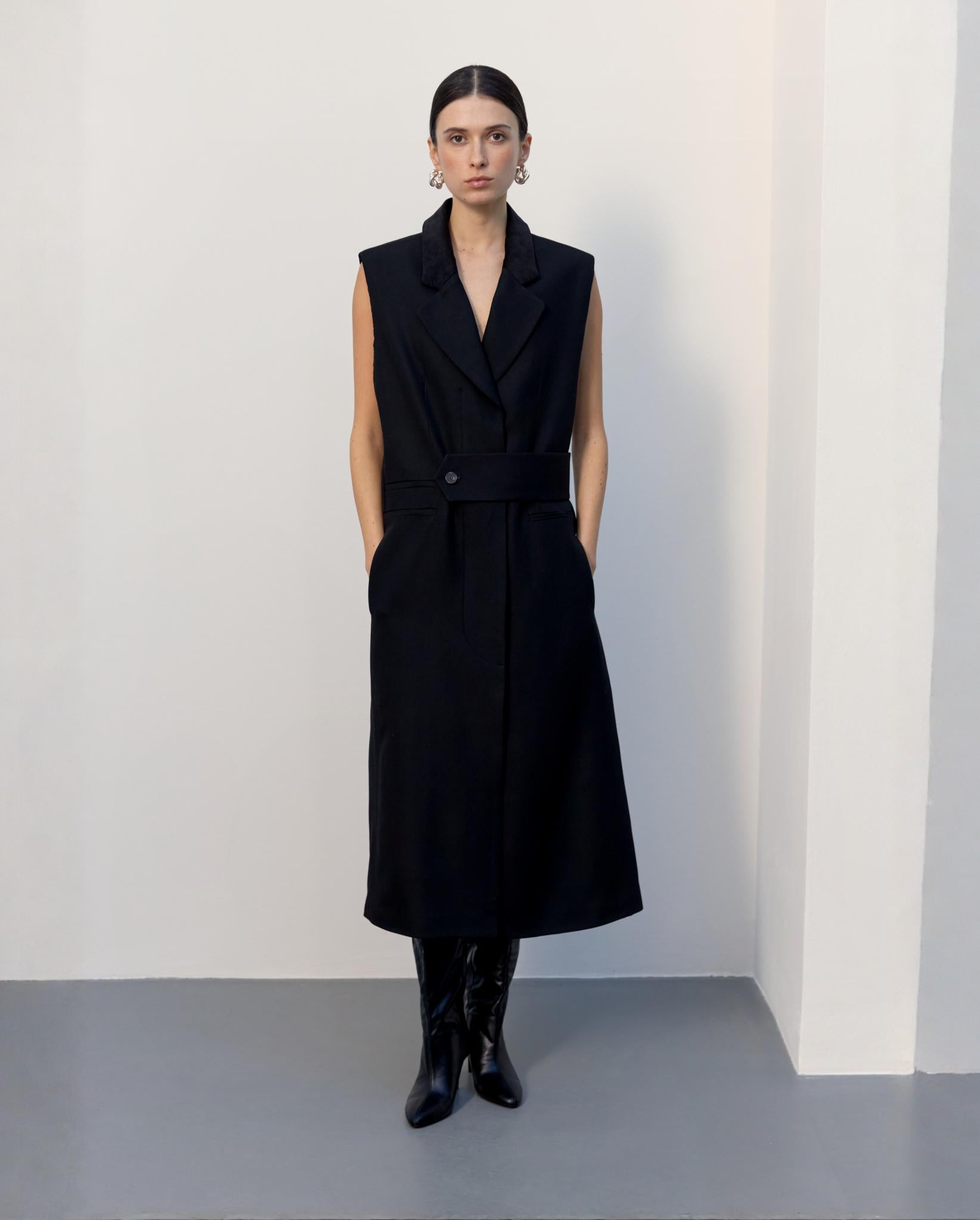 A woman wears the IVY OAK CORI ANN Vest, a sleeveless black trench coat dress with an adjustable belt, paired with black boots, standing against a plain white wall.