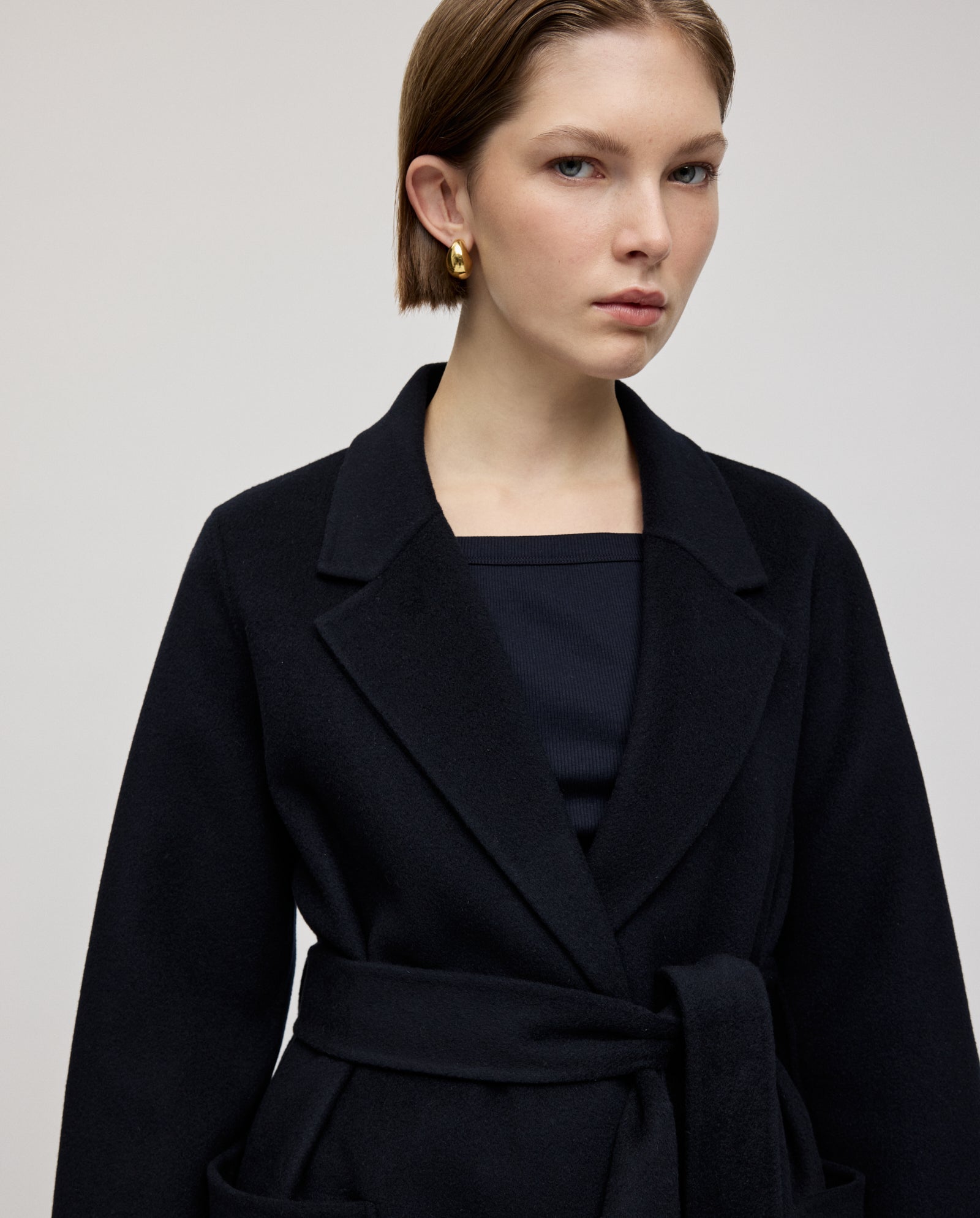 Woman wearing the IVY OAK CELIA Coat in black Midilänge with a belted waist and gold hoop earrings, looking at the camera with a neutral expression. The coat is crafted according to the Responsible Wool Standard.