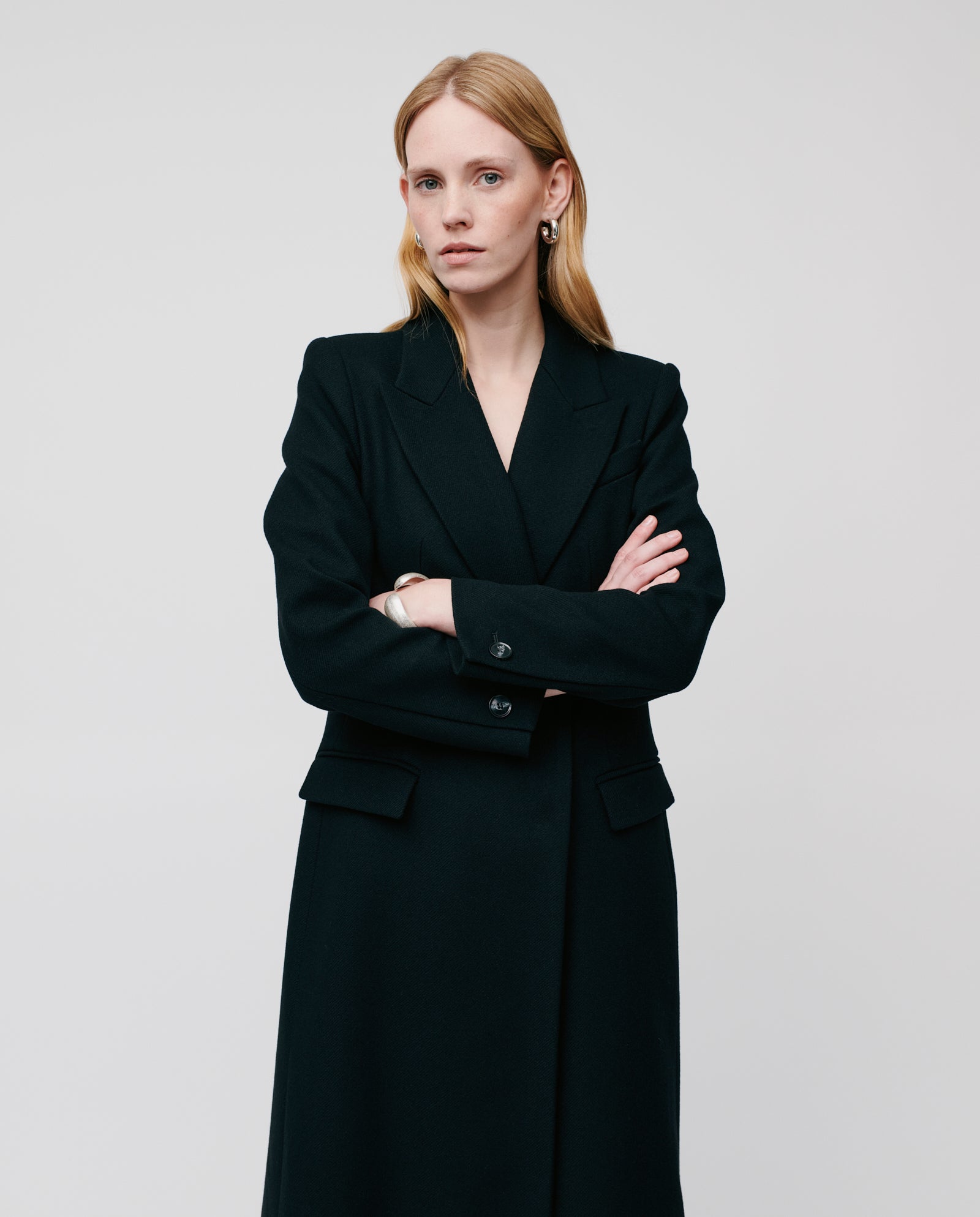 A woman stands with arms crossed against a plain, light background, wearing the IVY OAK CYNTHIA Coat crafted from luxurious Virgin Wool.