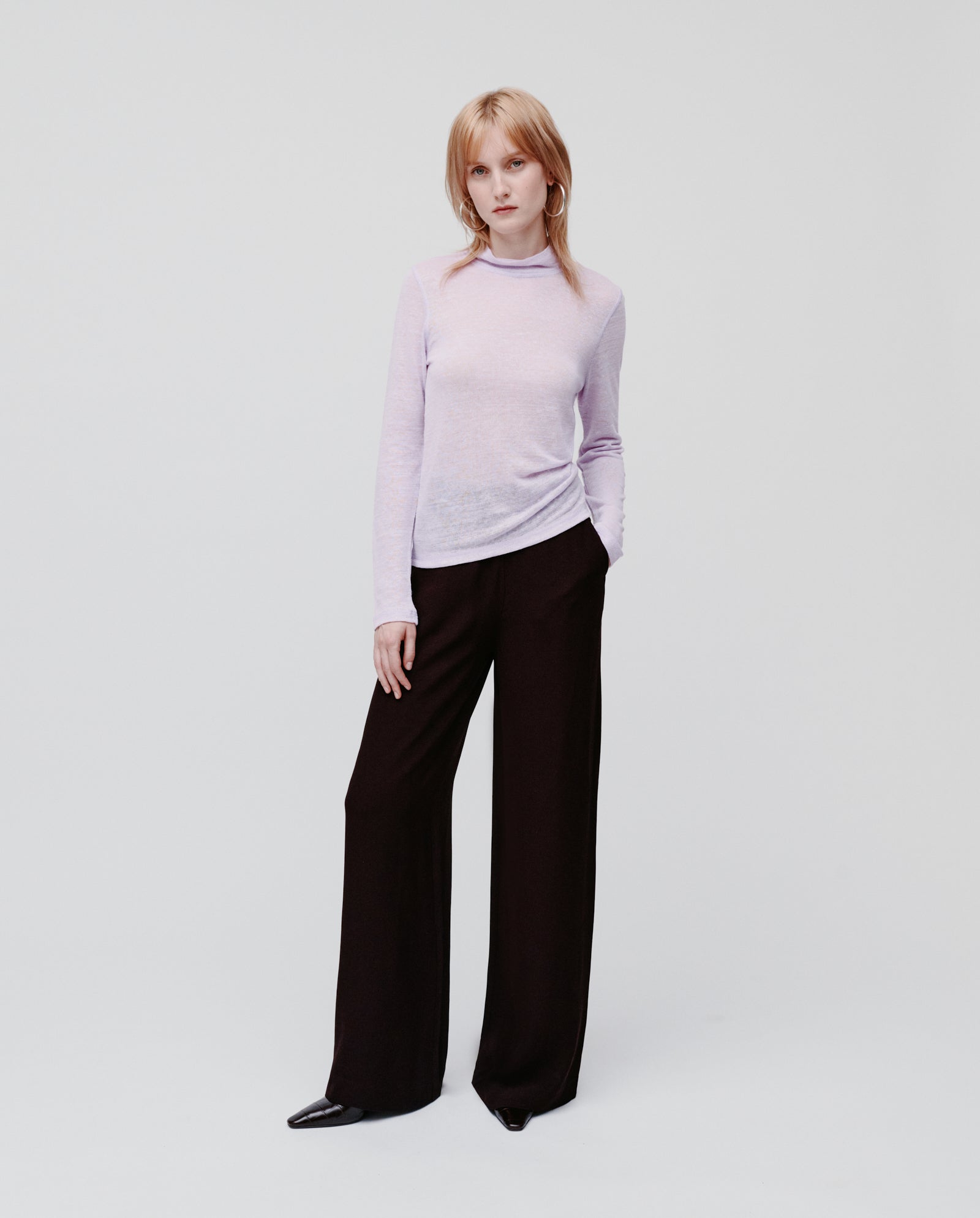 A woman wears the IVY OAK FELINE Top, a light purple thin jumper, paired with wide-leg black pants, standing against a plain white background.