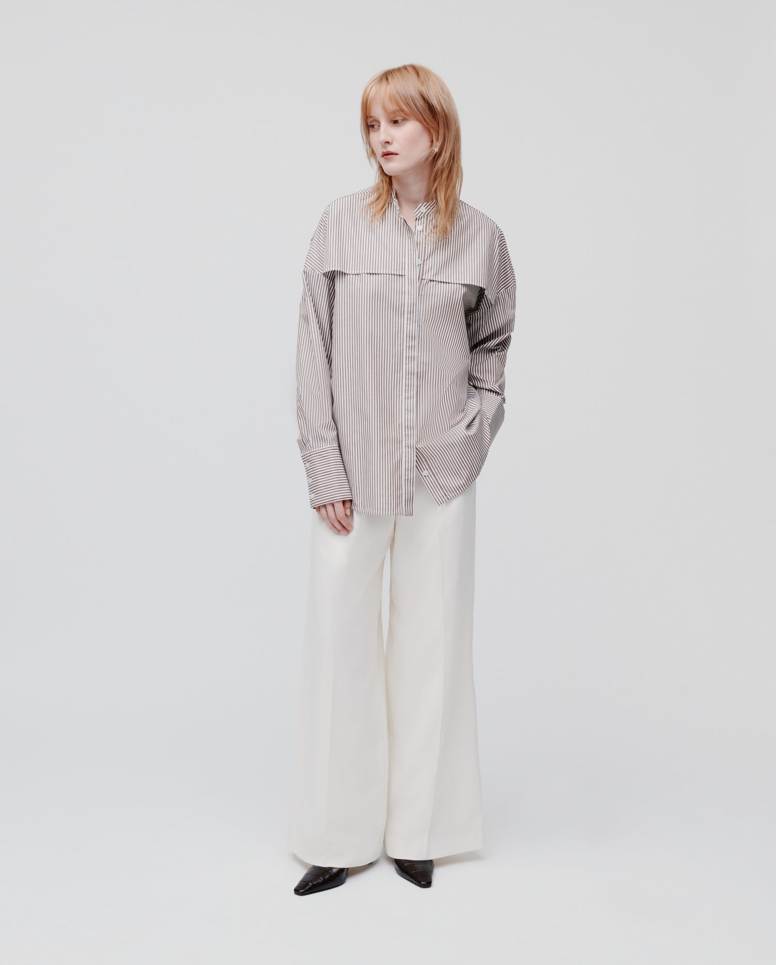 A woman stands against a plain white background wearing the IVY OAK EVE Blouse—an oversized striped shirt with a button closure—and wide white trousers.