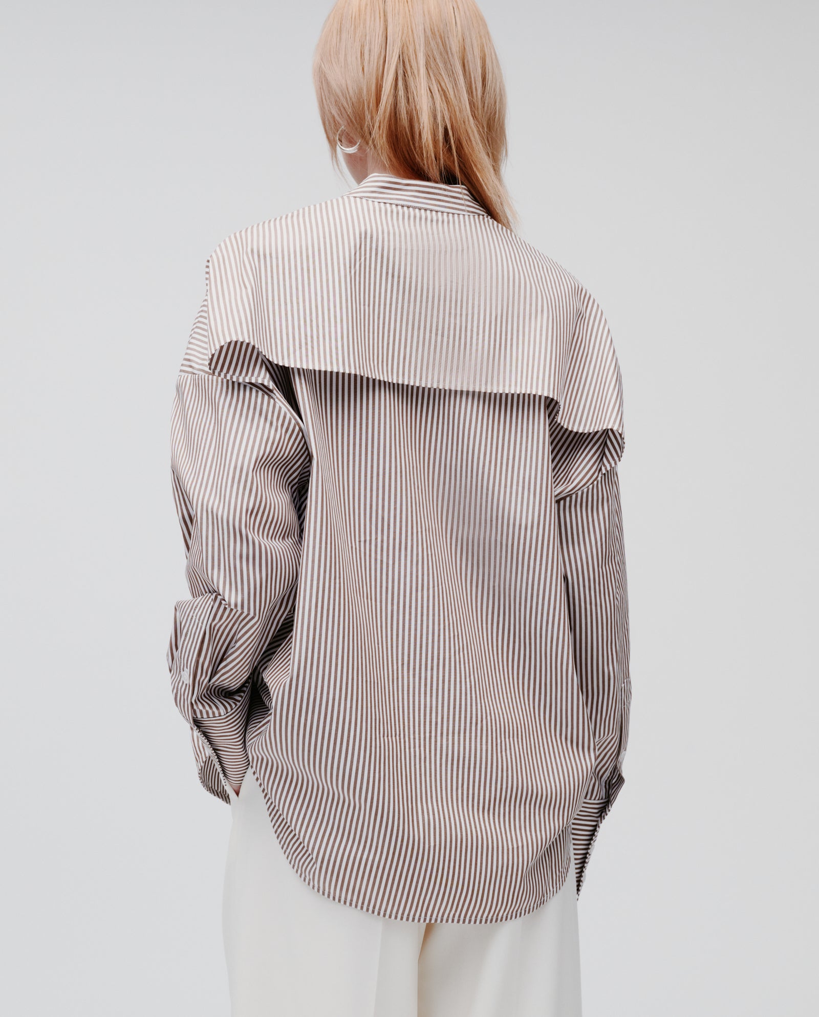 A person with long hair is shown from the back wearing the IVY OAK EVE Blouse—an oversized striped shirt with button closure—paired with white pants, against a plain background.