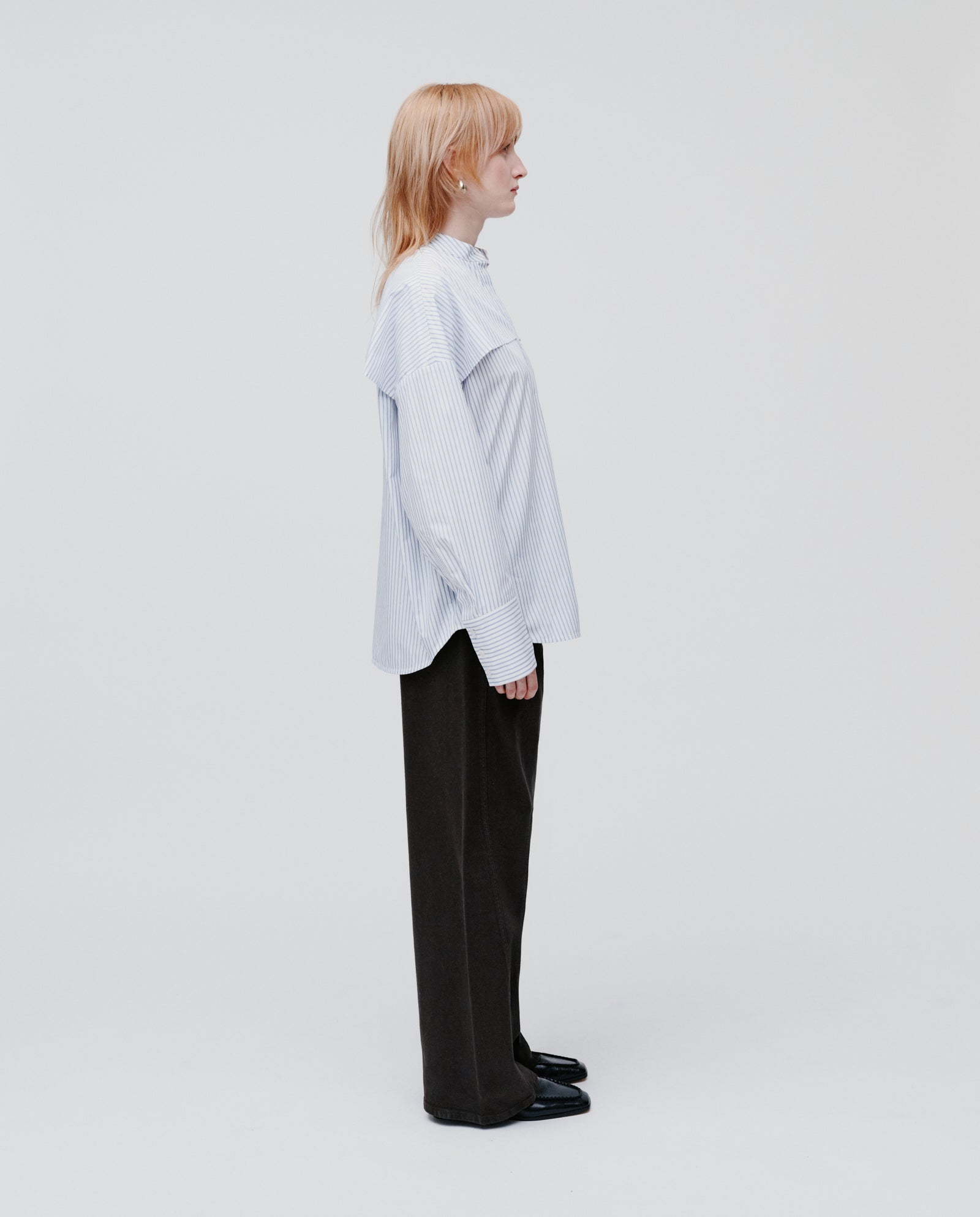 A person with light hair stands sideways against a plain white background, wearing the IVY OAK EVE Blouse—an oversized striped shirt with button closure—paired with dark pants.
